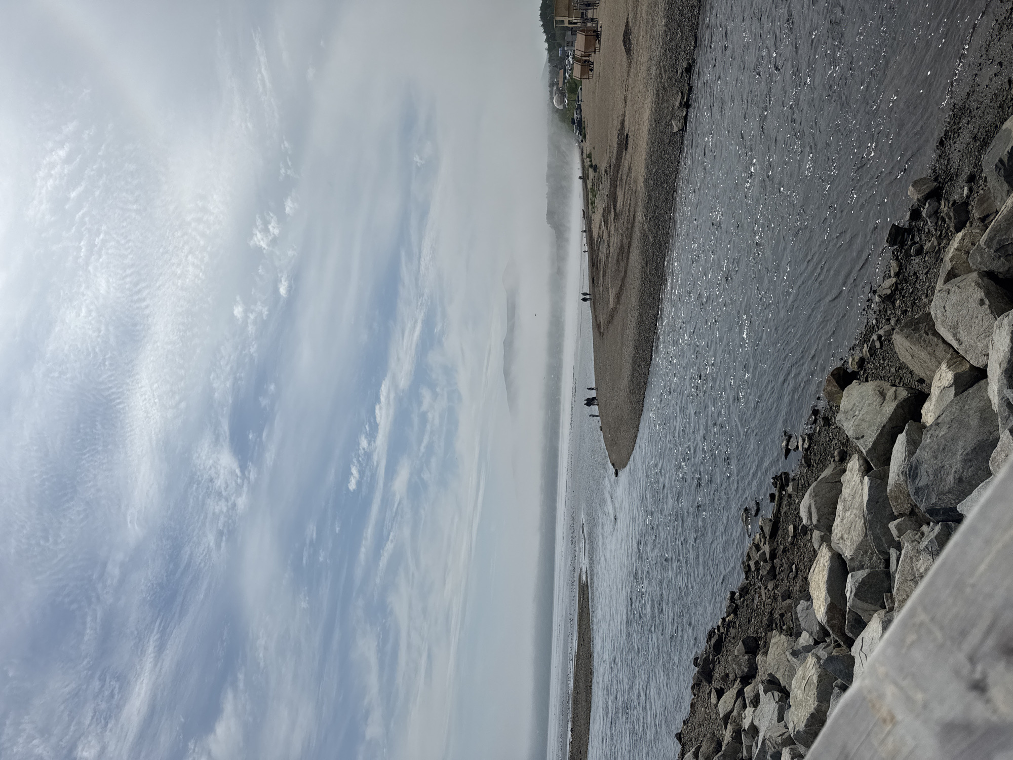 A wide-angle shot captures a serene coastal scene, likely in Magadan, Russia, under a hazy sky. In the foreground, a calm river flows from the right into a wider body of water on the left, its surface shimmering with reflected light. The riverbanks are lined with rough, grey rocks, some large and rounded, others smaller and more angular.
A narrow strip of sandy, pebble-strewn beach extends along the right side of the river. A few small groups of people, appearing as distant figures, are scattered along this beach, some walking, others standing. Beyond the beach, the hazy atmosphere obscures the distant landscape, revealing rolling hills or mountains veiled in a soft, white mist. A few structures, possibly buildings and tents, are visible on the far right of the beach, along with several parked vehicles.
The sky above is a pale blue, streaked with wispy, cirrus clouds that form delicate patterns across the expanse. The overall impression is one of quiet contemplation and the natural beauty of a coastal environment.
