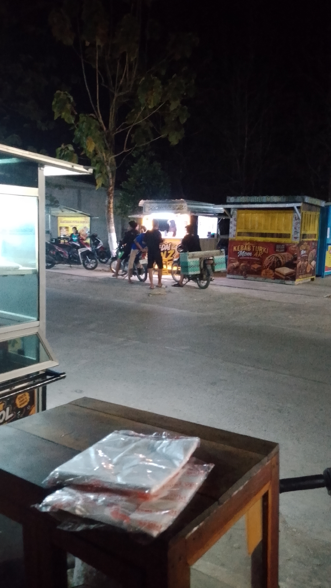 The image depicts a nighttime scene at a street food stall area, possibly in Karanglangu, Indonesia. In the foreground, a wooden table holds two stacks of items wrapped in clear plastic. The background features several food stalls with bright lighting, suggesting commercial activity.
One stall has a sign with the text "KEBAB TURKI Mom AR," and displays images of kebabs and other food items. Another stall appears to be a general food vendor, with people visible inside. Several motorcycles are parked around the stalls, indicating transportation used by patrons or vendors.
A group of individuals, likely customers and vendors, are gathered around the stalls. Some are standing and conversing, while others appear to be ordering or waiting for food. The overall ambiance is that of a bustling night market or street food hub. The presence of the tree and the dark sky suggest an outdoor location.