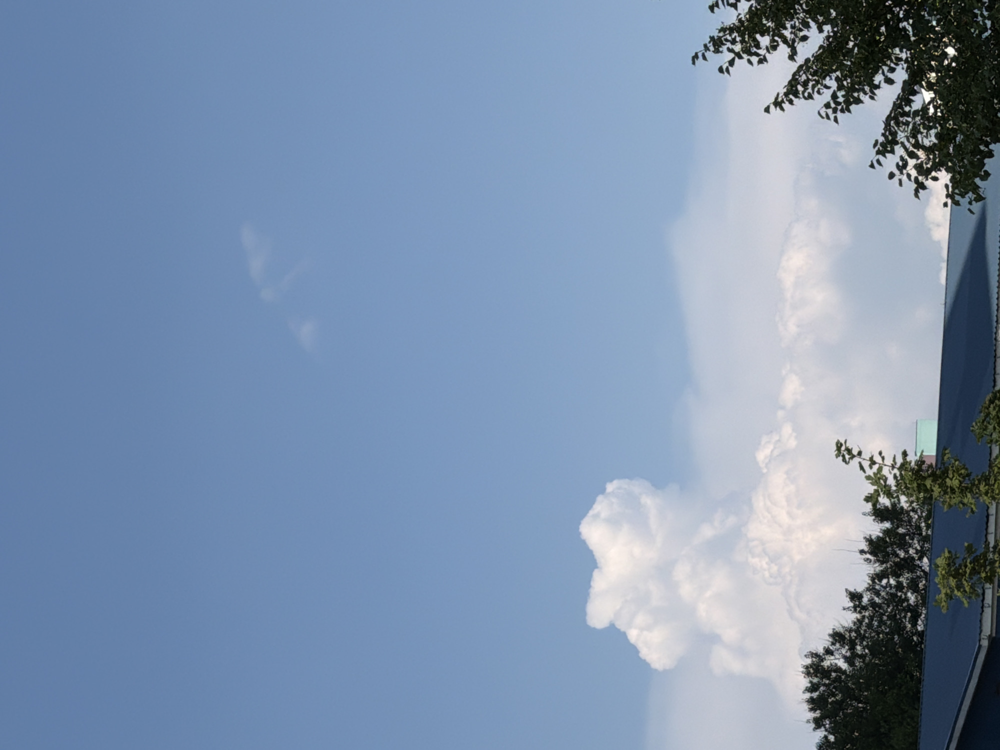 The image shows a bright, clear blue sky with some wispy clouds. In the lower portion of the image, a large, fluffy cumulus cloud formation is visible. Below the clouds, the tops of dark green trees and the roof of a building with a blue-gray surface are discernible. A portion of a light blue and pink building is also visible behind the trees. The scene appears to be an outdoor, daytime view. The specific location in Kyiv, Ukraine cannot be determined from the visual information provided. There are no people or discernible actions in the image.
