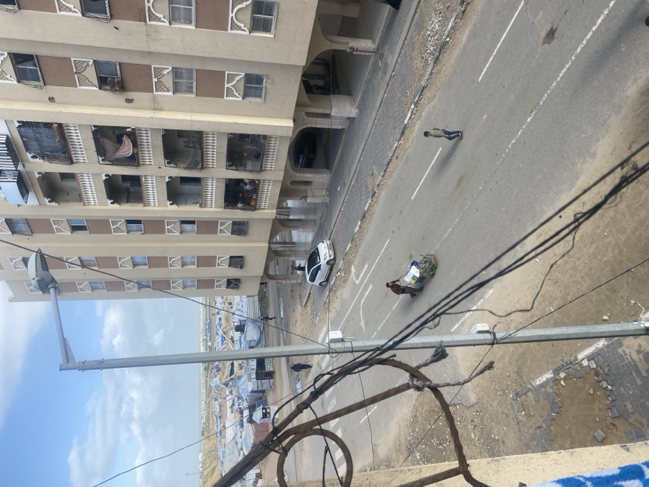 The image depicts an elevated view of a street scene in Khan Yunis, Palestinian Territory, under a partly cloudy sky.
In the immediate foreground, part of a light-colored building wall is visible on the left, with tangled electrical wires and a gray metal streetlight pole obscuring the lower-left portion of the frame. A blue and white patterned fabric hangs over an unseen structure on the far left.
The midground features an asphalt road with white solid and broken lane markings. An adult male, dressed in a dark red shirt and dark trousers, pushes a green two-wheeled handcart loaded with white and blue bundled materials along the right side of the road, moving away from the viewer. Further down the road, another individual, possibly a young male, in a light-colored top and dark trousers, walks in the middle of the lane, also moving away from the viewer. A white compact car is parked partially on the dirt shoulder next to the sidewalk on the right, facing the viewer. Another vehicle, possibly red, is partially visible under the arches of the large building on the right. Paved sidewalks run along both sides of the road, with adjacent dirt and gravel patches.
The background on the right is dominated by a multi-story, light-colored building with brown vertical accents and decorative arch-shaped window frames. It features multiple balconies with white railings, some of which have laundry or fabrics hanging. Several individuals are visible on the balconies and through windows. The ground floor consists of open archways suggesting covered parking or passage. To the left, beyond the road and a dirt expanse, a large, dense encampment stretches into the distance, characterized by numerous makeshift shelters and tents, many covered with blue tarpaulins, extending towards a flat horizon.