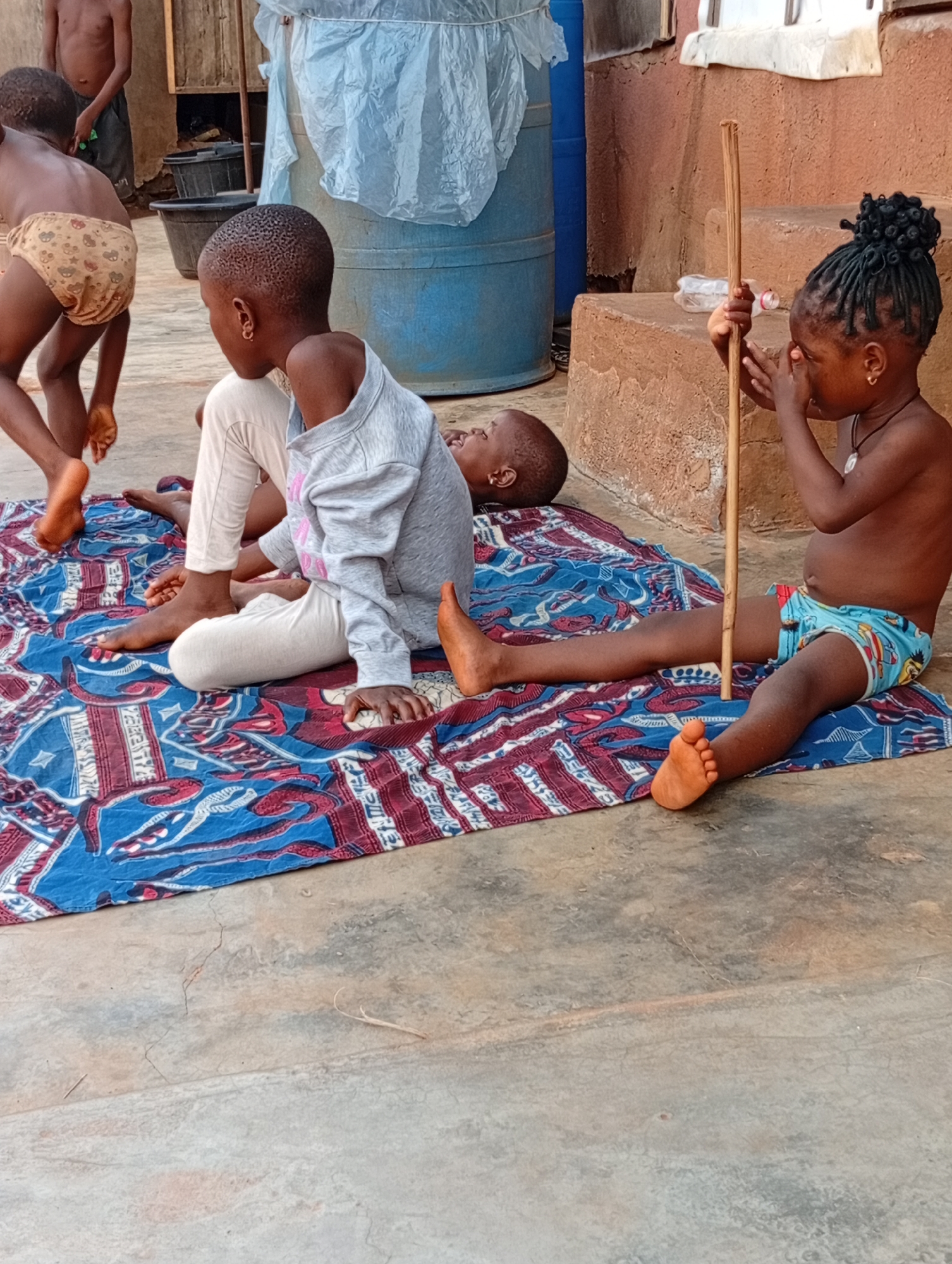This outdoor image depicts several children in a casual setting, likely a residential yard in Ogidi, Nigeria. The ground is a light grey concrete, and a colorful mat with red, blue, and white patterns is spread across a portion of it.
On the far left, a child wearing light-colored, patterned briefs stands with their back to the viewer, their bare feet visible. Another child or adult is partially visible further in the background to the left. On the patterned mat, a child in a light grey long-sleeved top and light-colored trousers sits, facing away from the camera, with their bare feet on the mat. Another child is lying down next to them, mostly out of view, with just a portion of their head visible.
To the right, a young child sits on the mat, shirtless and wearing light blue patterned briefs with cartoon characters. This child has dark braided hair styled into multiple small buns on top of their head, and is wearing a single earring and a necklace with a small pendant. They hold a long, thin wooden stick upright with both hands, with their fingers partially covering their eyes, as if playing or looking through them.
In the background, against a reddish-brown concrete or mud-rendered structure, two blue plastic drums are visible, partially covered by a translucent white plastic sheet. Several black plastic buckets are also present near the drums. An empty, clear plastic bottle rests on the steps of the reddish-brown structure.
The scene takes place during the daytime under natural light. The children are barefoot or lightly dressed, suggesting warm weather. No visible text is present in the image.