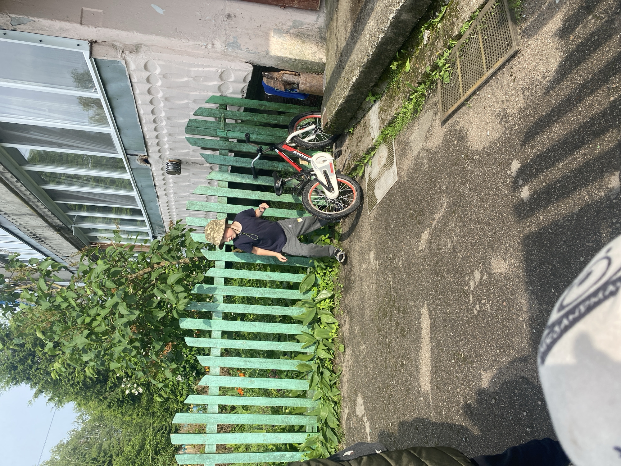 This image depicts an outdoor scene in Ihnatpil, Ukraine, featuring a young boy, a bicycle, and a residential building under natural light, casting prominent shadows.
In the mid-ground, a young boy, approximately 6-8 years old, is visible. He wears a camouflage bucket hat, a dark t-shirt, gray sweatpants, and dark sandals. He is positioned slightly left of center, with his left leg raised and right foot on the ground, indicating movement. He looks towards the right side of the frame.
To the boy's right, a red, black, and white children's bicycle, branded "ARDIS" on its frame, is parked on a concrete path, leaning against a light green wooden picket fence. The bicycle has red rims. The fence extends across the middle of the frame, bordering an overgrown garden area with various green plants and some orange flowers.
Behind the fence, a lush green bush with dark leaves is prominent, with taller trees and a brighter sky in the background. To the right of the fence, a multi-story residential building is visible. Its facade is a faded pinkish-beige with a textured pattern of circular indentations on the lower wall. Above this, a long, enclosed balcony or series of windows with white frames extends across the building. Below the windows, a metal vent or pipe fixture protrudes from the wall. Near the ground level, to the right of the bicycle, a small, dark rectangular opening is present in the building's foundation, with a wooden log or stump and a rusty metal barrel nearby. A blue cloth hangs near this area.
The foreground consists of a gray, asphalt-like concrete ground surface. Long, striped shadows from the fence pickets are cast onto the ground, indicating direct sunlight from the left or right. In the bottom left corner, part of a white and blue football with the word "SANTANA" partially visible is in extremely close foreground, suggesting its proximity to the camera or observer. Two metal drainage grates are present on the ground: a smaller one to the left and a larger, rectangular one to the right, adjacent to a raised concrete curb or ramp that runs along the right edge of the path, bordered by weeds and grass.