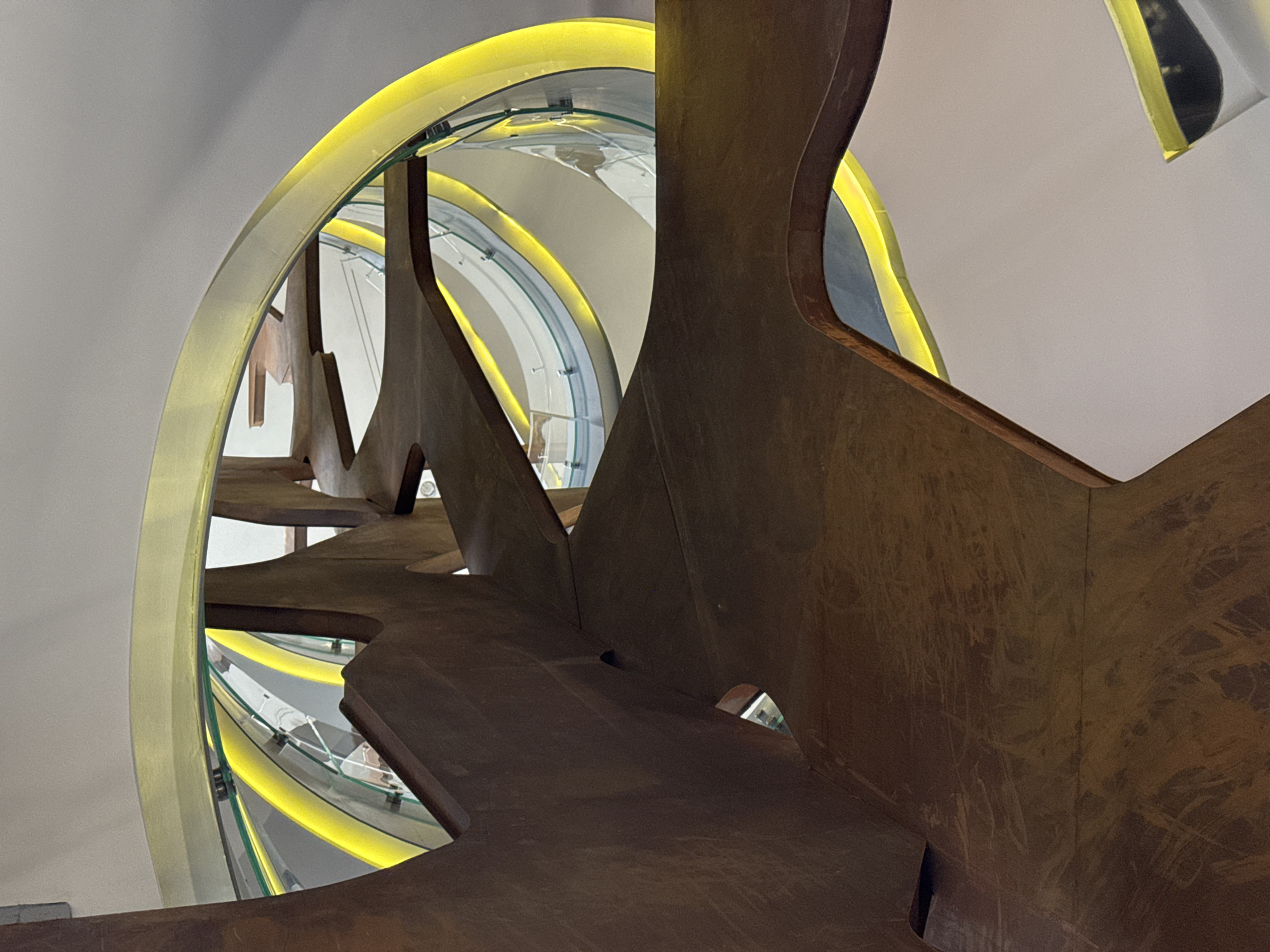 The image shows a close-up, low-angle view of the interior of a spiral staircase, likely in a modern building. The main elements are the weathered, rusty-brown metal structure of the staircase, which appears to be made of thick, curved panels with sharp, almost organic cutouts. These panels form the steps and railings.
Around the central void of the staircase, a curved glass railing is visible, illuminated by a continuous strip of bright yellow light embedded along its top edge. This yellow light reflects off the polished surfaces of the staircase and the glass, creating a dynamic interplay of light and shadow. The background shows the curving walls and floors of the upper levels of the building, also illuminated by the yellow light, giving a sense of depth and height. The overall impression is one of a contemporary architectural design with an artistic, almost sculptural quality.
This scene is located in the atrium of the CaixaForum Madrid, an art and cultural center designed by architects Herzog & de Meuron. The building is renowned for its distinctive vertical garden facade and its striking interior atrium.