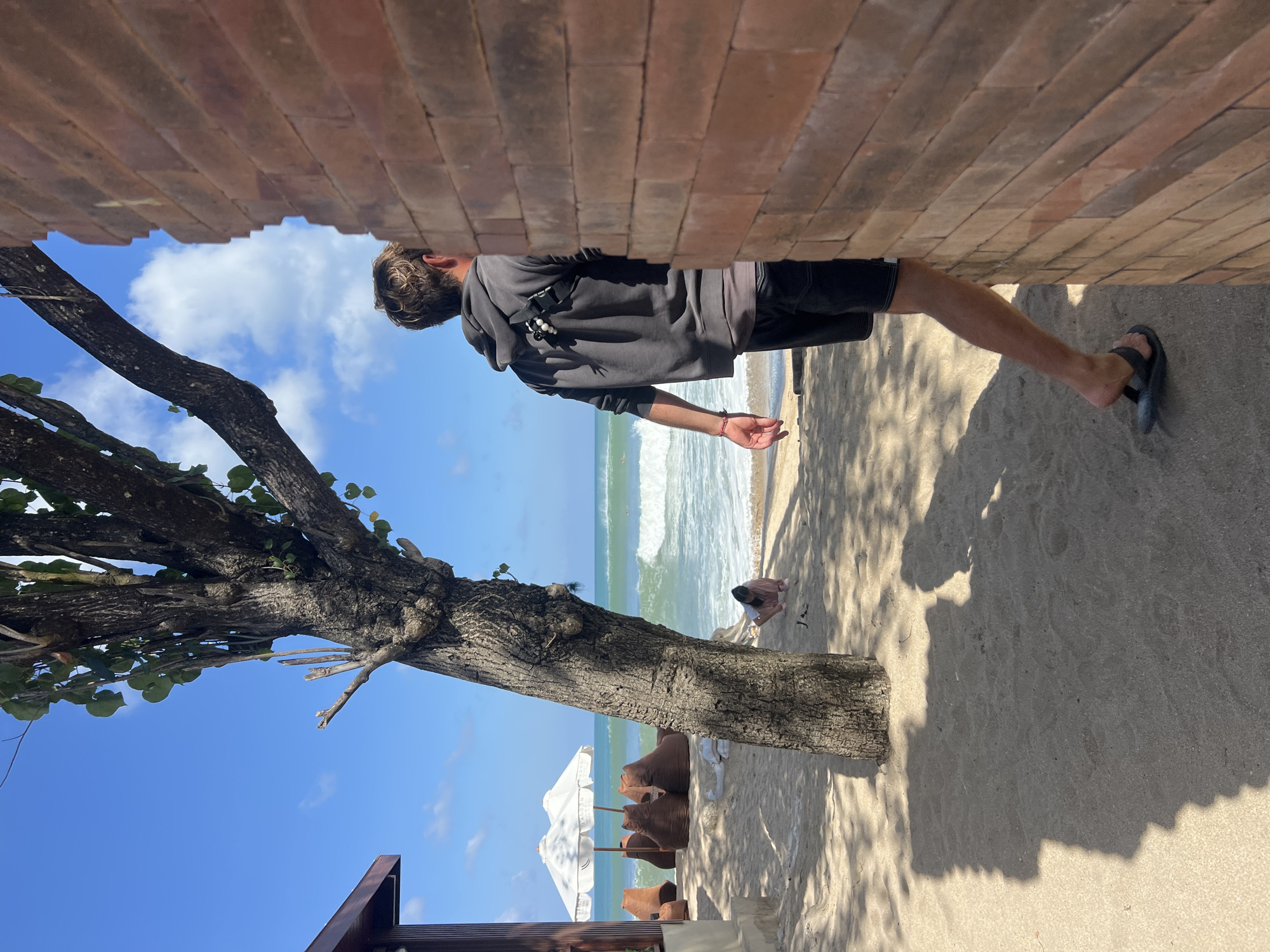 A man with blonde hair wearing a grey hooded sweatshirt and black shorts is standing on a sandy beach, facing away from the camera and looking towards the ocean. He is partially obscured by a brick wall on the right side of the frame. The beach has a tree with a thick trunk and sparse leaves in the foreground, casting a shadow on the sand. In the background, there are two white beach umbrellas and several brown beanbag chairs. The ocean has light green waves and a clear blue sky with some clouds overhead. A person is sitting on the sand near the water's edge in the distance. This scene is located on the beach in Kuta, Indonesia.