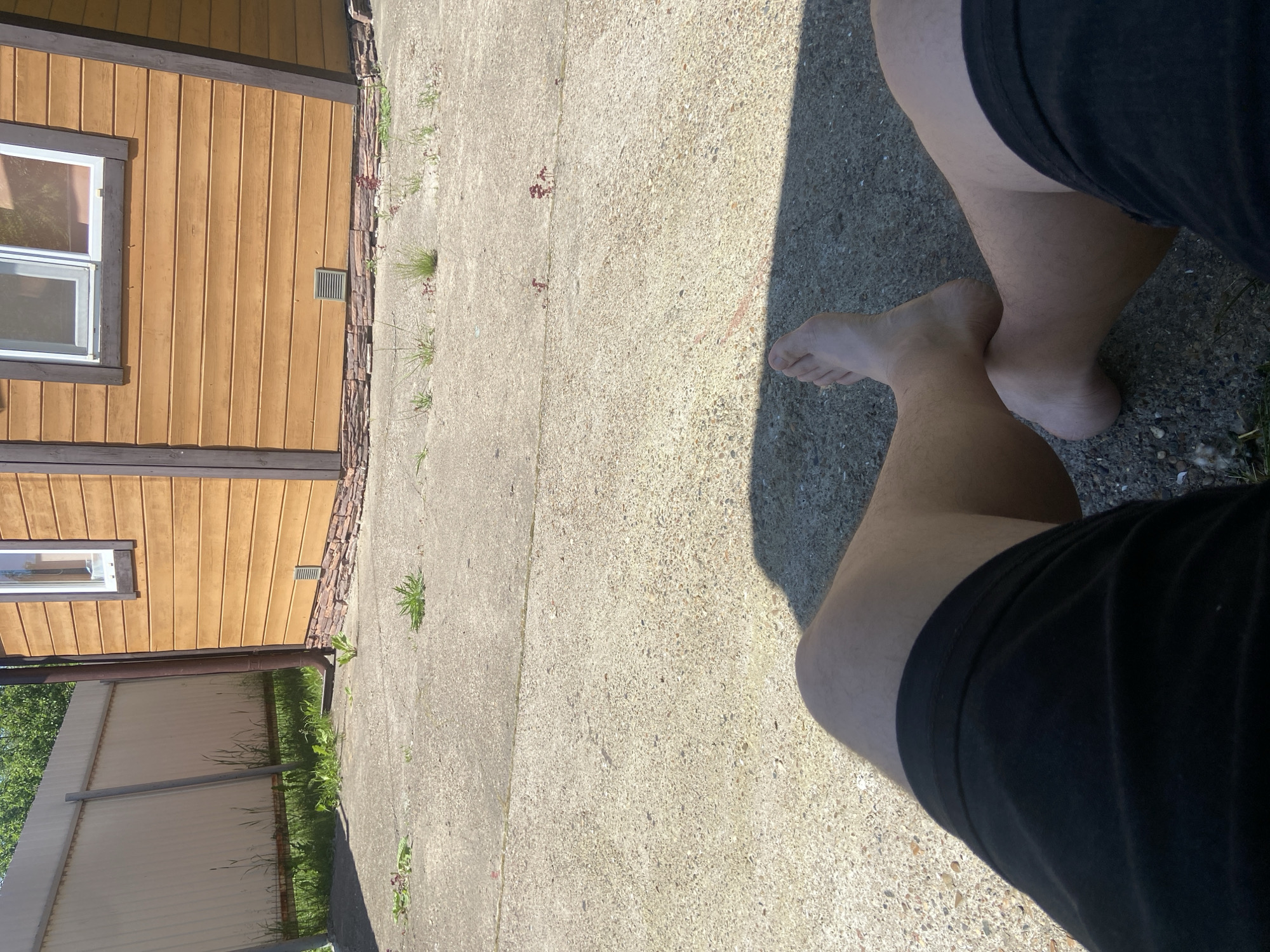 A first-person perspective shot shows a person sitting on a concrete patio in front of a small, rustic wooden building. The building has a brown, wood-paneled exterior with a single window featuring white trim. A dark brown downspout runs along the side of the building, emptying onto the concrete. The patio is cracked in places, with sparse grass and weeds growing through the fissures. A light-colored metal fence runs along the left side of the scene, with greenery visible behind it. The person's legs, clad in black shorts, are crossed at the ankles, and their bare feet are visible. The scene is illuminated by bright sunlight, casting a shadow on the patio. The location is identified as Kazan, Russia.