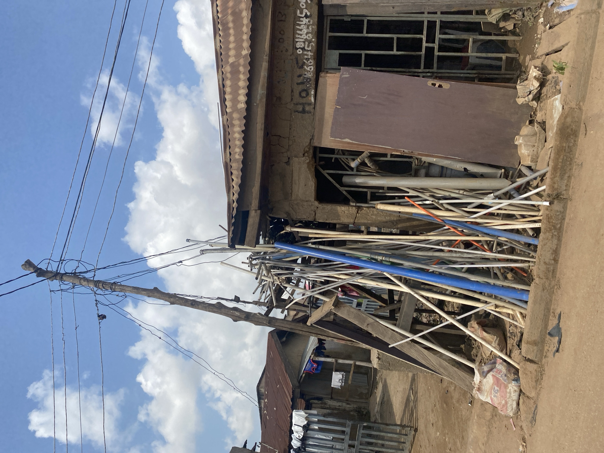This is a forensic, matter-of-fact description of a scene in Ado, Nigeria.
The image depicts an outdoor environment, possibly a street or alleyway, with signs of an urban setting in Ado, Nigeria. The foreground is dominated by a dirt or unpaved road surface. On the right side of the image, a building with a corrugated metal roof is visible. The walls of this building appear to be constructed from rough, possibly mud or unrendered concrete blocks. Scrawled text, including phone numbers like "08166645858", is visible on the building's wall. In front of this building, a large, flat, brown wooden object, resembling a door, leans against the structure. To the left of the door, a metal grate or window covering is present.
A significant accumulation of discarded materials, predominantly pipes of various sizes and colors (including blue and white), along with wooden planks and metal rods, is piled in front of the building and extending towards the left. Some of these pipes are leaning against a roughly constructed wooden frame. A large, multi-colored sack, possibly containing cement or building materials, lies on the ground to the left of the pipes.
Further to the left, another building with a rusted corrugated metal roof is partially visible. Laundry, including white garments, is hanging on a line in front of this building, and a gate made of metal bars is also present.
Overhead, a prominent, rough-hewn wooden pole stands tall, serving as a makeshift utility pole. Numerous electrical wires are haphazardly strung from this pole, creating a tangled network against a bright blue sky with scattered white clouds.
The scene conveys a sense of disarray and perhaps a low-income or informal commercial area. There are no people clearly visible in the image. The overall atmosphere is that of a functional but untidy urban space.