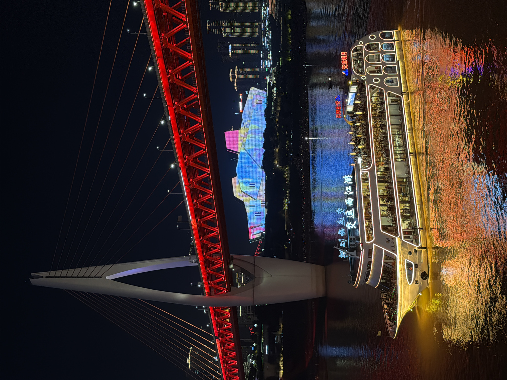 This is a night-time, long-shot, eye-level, outdoor photograph of a wide river with a large, multi-decked tour boat illuminated with white and yellow lights, cruising on the water. The boat, a prominent subject, is centered in the lower half of the frame, moving from left to right. Its reflection shimmers on the dark water, tinged with orange and yellow from the boat's lights.
Behind the boat, in the middle ground, a massive, modern suspension bridge is visible, illuminated with bright red lights. The bridge's white pylon, on the left side of the frame, reaches high into the dark night sky, supporting the red structure with numerous cables. Further in the background, beyond the bridge, a cityscape of illuminated skyscrapers rises. To the right of the bridge pylon and in front of the skyscrapers, a large, strikingly illuminated building displays a vibrant, multi-colored light show, with hues of blue, pink, and green. The overall scene suggests a lively urban waterfront at night, possibly in the Yuzhong District of Chongqing, China, given the distinctive architecture of the bridge and the illuminated buildings characteristic of this area. The dark sky is devoid of stars but has a slight gradient, indicating urban light pollution.