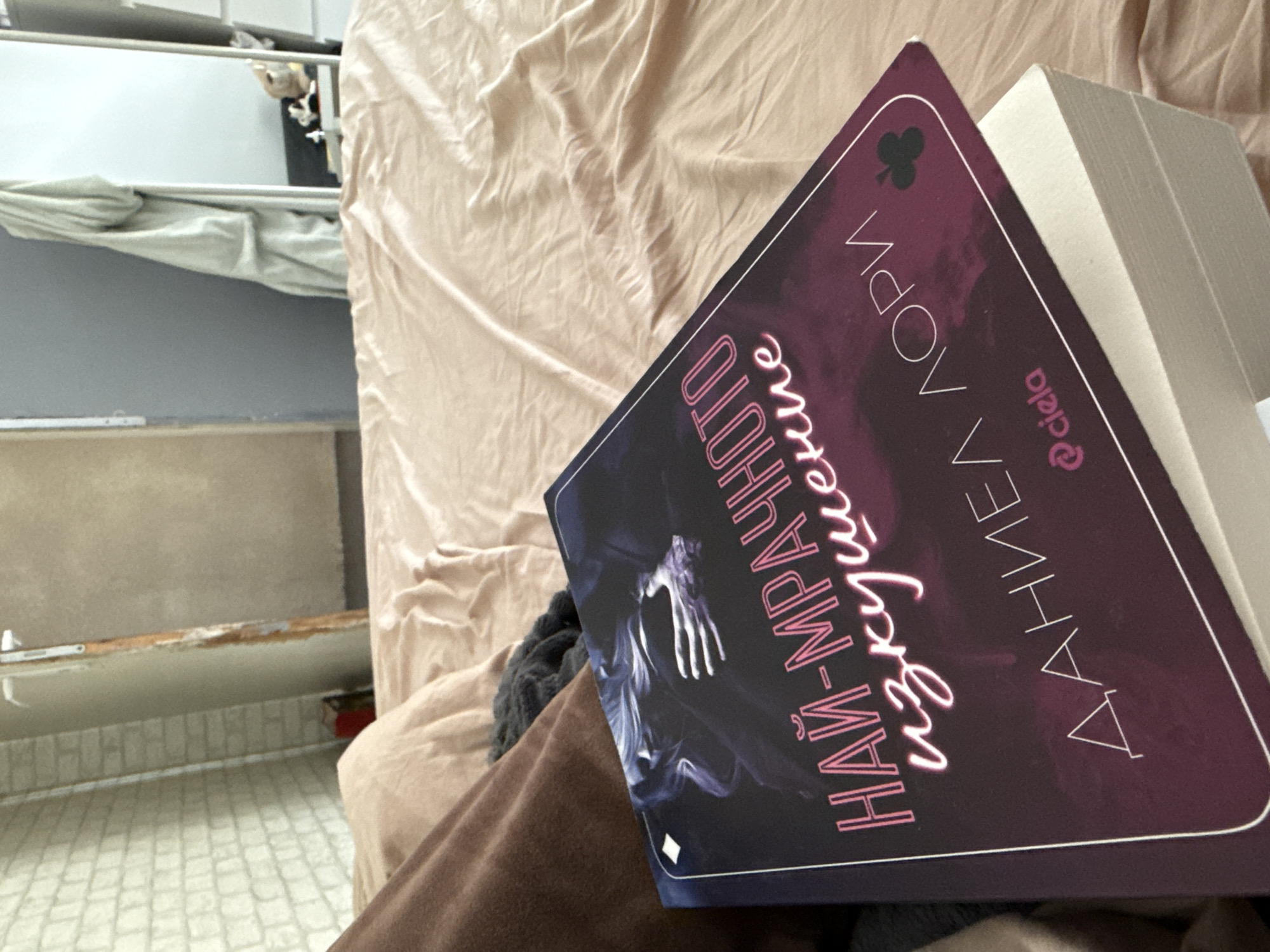 The image displays a close-up, downward-angled view of a book resting on a person's lap or leg, indicated by a dark brown fabric. The book's dark-themed cover features a white border and a prominent club suit symbol in the top left corner. The title, in Cyrillic pink outline font, reads "НАЙ-МРАЧНОТО изкушение" (The Darkest Temptation). The author's name, also in Cyrillic and rotated, is "ДАНИЕЛ АЛОРИ" (Danielle Lori), with "@ciela" visible as the publisher or imprint. A skeletal hand design is part of the book's cover art.
The book is positioned against a background of wrinkled, light beige bedding, suggesting it is on a bed. In the left background, a metal bunk bed frame is visible, with a light grey sheet or fabric draped over the upper rail. A light grey wall runs below the bunk bed, meeting a light-colored, small rectangular tiled floor. The scene depicts a moment of reading or a pause from it. The indoor setting, with a bunk bed and simple decor, suggests a bedroom or dormitory environment. The Cyrillic text on the book is consistent with the provided location context of Sofia, Bulgaria. The lighting is ambient, characteristic of indoor conditions.