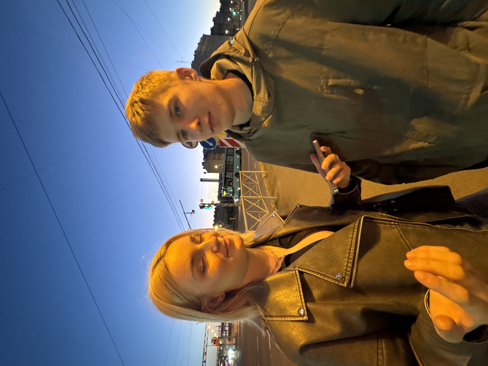 The image depicts a young man and a young woman standing on a sidewalk at dusk in Saint Petersburg, Russia. The sky is a deep blue, indicating either early morning or late evening. The woman, on the left, has blonde hair and is wearing a black leather jacket over a dark top. She is holding a smartphone in her right hand, gesturing with her left hand. The man, on the right, has short blonde hair and is wearing a hooded olive-green jacket. He is holding a smartphone in his left hand and appears to be looking off to the side. In the background, a street with traffic lights, streetlights, buildings, and a bus is visible, suggesting an urban environment. A metal fence runs along the sidewalk. The lighting suggests artificial streetlights are illuminated.