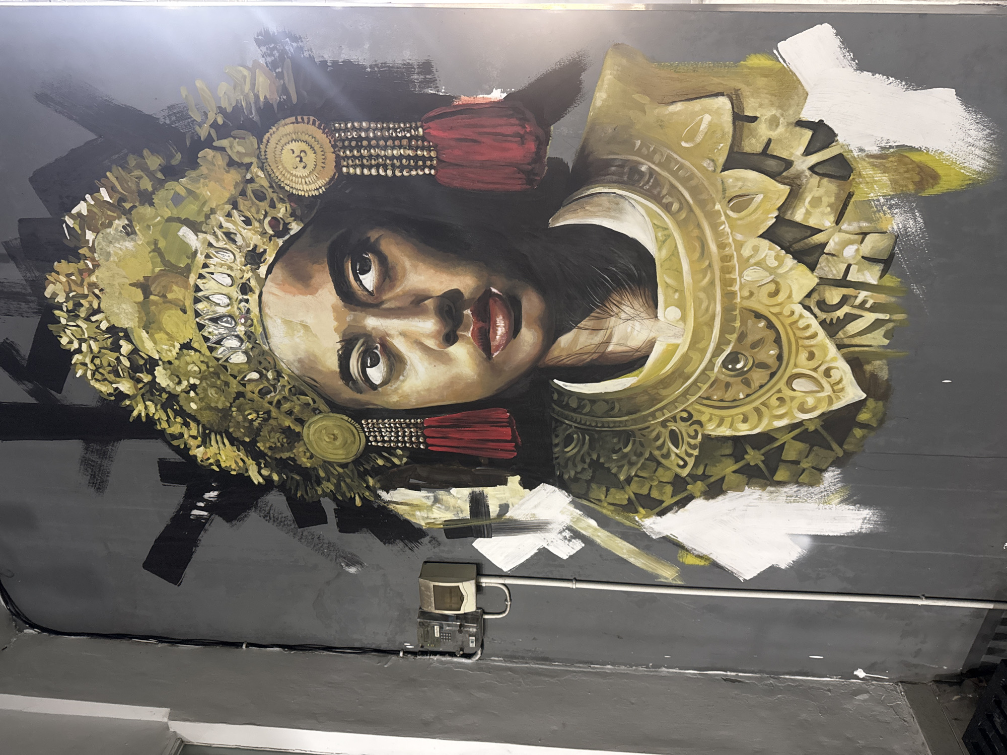 This is a photograph of a ceiling mural depicting a portrait of a woman with elaborate headdress and ornamentation, likely in a Balinese style given the location context of Ubud District, Indonesia. The woman's face is rendered in a realistic, yet somewhat stylized manner, with her gaze directed upwards and slightly to the left. She wears a large, golden headdress adorned with intricate patterns, beads, and red tassels. Her neck and shoulders are also decorated with ornate gold jewelry. The background of the mural is a dark, textured gray, with bold brushstrokes of black, gold, and white paint suggesting a dynamic and perhaps unfinished quality to the artwork.
Below the mural, a metal conduit runs horizontally along the wall, attached to a small, light-colored electrical box or meter. The overall impression is that of a vibrant, culturally inspired artwork displayed in an indoor setting, possibly a public space or a building interior in Ubud. The lighting appears to be artificial, and the image is taken from a low angle, looking up at the ceiling. There is no visible text in the image.