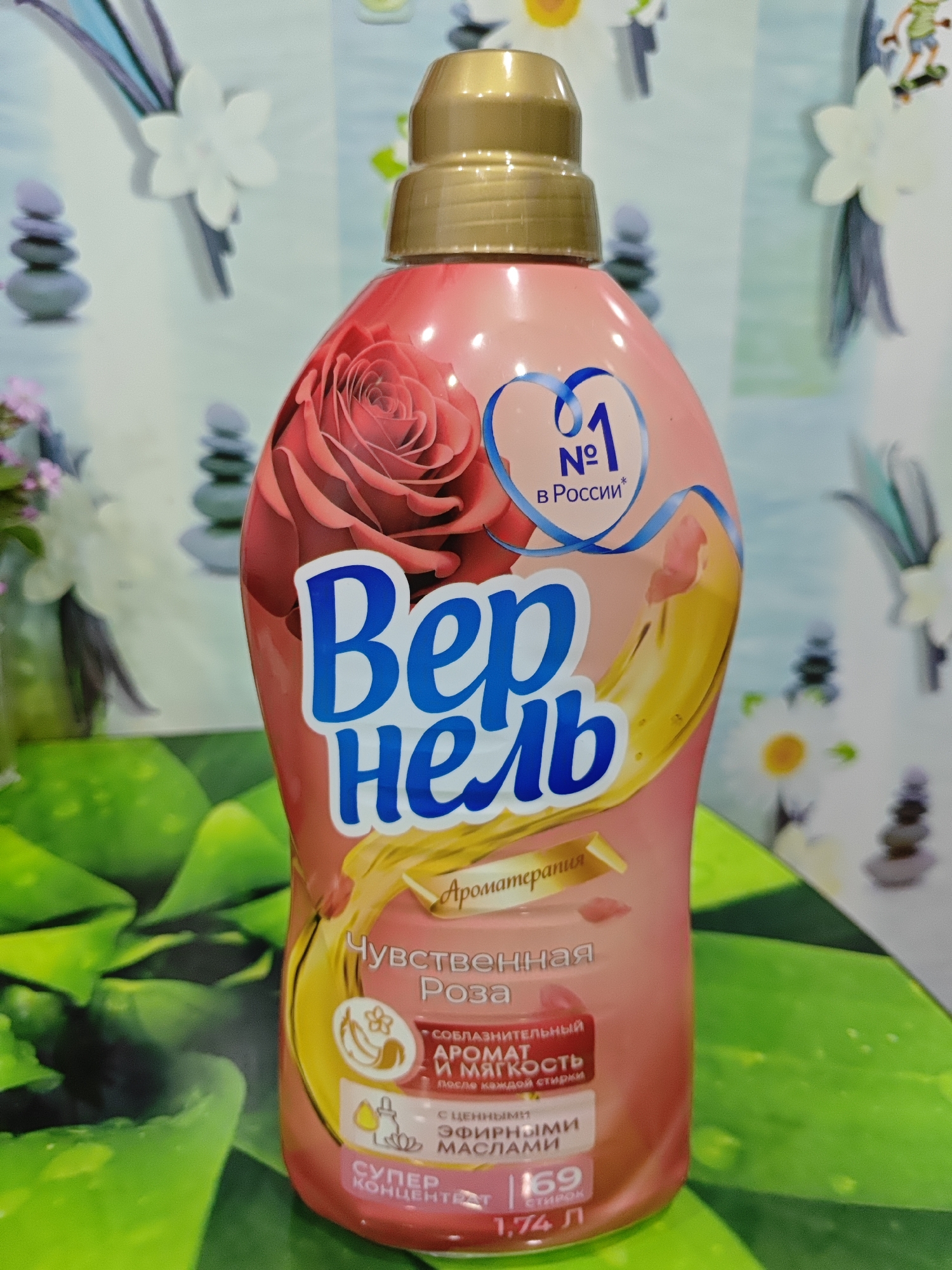 A pink plastic bottle of fabric softener, labeled "Вернель" (Vernell) and "Аромат Розы" (Rose Aroma), is displayed against a blurry background. The bottle features a large red rose illustration and a blue heart with the number "1" inside, signifying "№1 в России" (No. 1 in Russia). Below the main text, there are smaller labels indicating "Чувственная Роза" (Sensual Rose), "Смягчительный аромат и мягкость после каждой стирки" (Softening aroma and softness after each wash), "с ценными эфирными маслами" (with valuable essential oils), and "СУПЕР КОНЦЕНТРАТ 1,74 л 169 стирок" (SUPER CONCENTRATE 1.74 L 169 washes). The bottle is positioned on a green surface with a patterned texture, possibly a tablecloth or placemat. The background is out of focus and appears to be a wall with decorative elements, including stacked stones, flowers, and abstract shapes. There are no people visible in the image. The scene is indoors, likely in a home environment. The location of Polyarny, Russia, is not visually evident from the image.