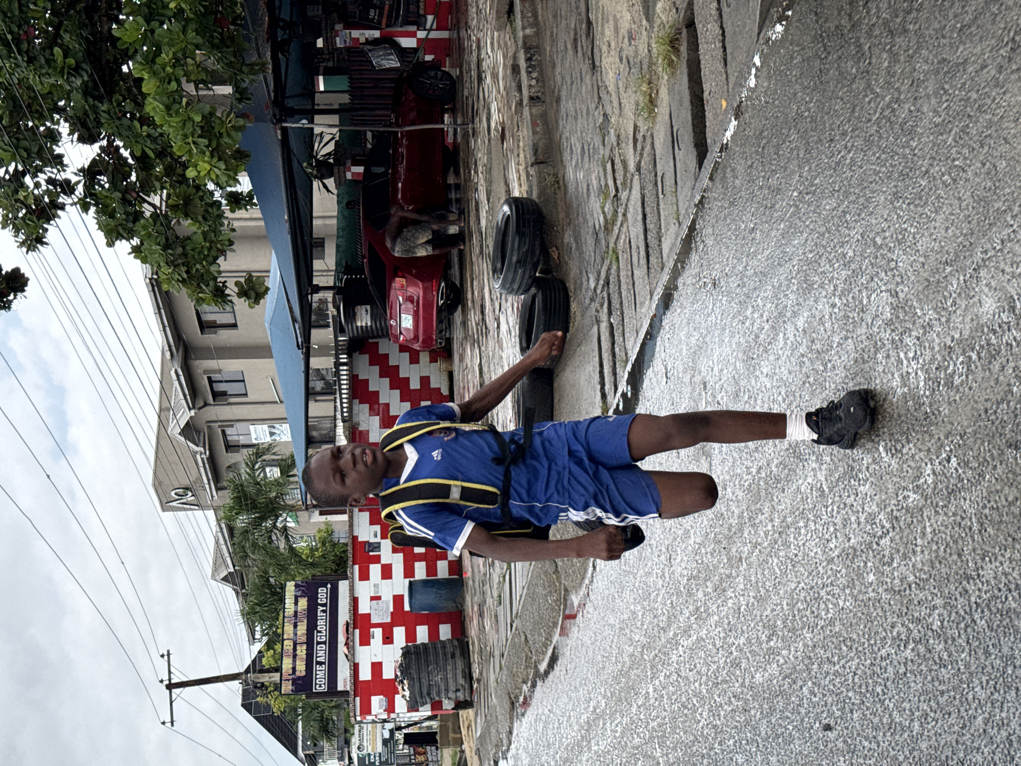 A young male, wearing a blue and white Adidas soccer uniform with a yellow and black harness, is walking on a wet sidewalk in Ado, Nigeria. The sidewalk shows signs of recent rain. Behind him is a street with a building featuring a red and white checkered design and signage for "THE PROMISED BRETHREN CHURCH WORLDWIDE. COME AND GLORIFY GOD." A red car and some tires are visible near the building. Overhead, power lines crisscross the sky near trees.