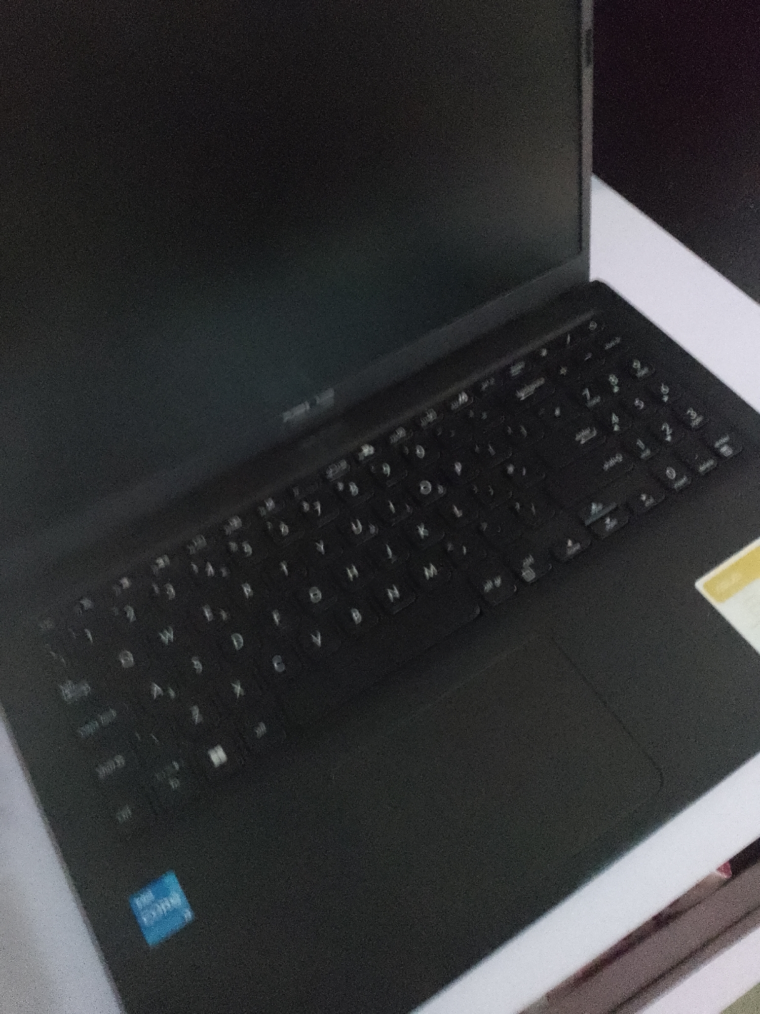 The image displays a close-up, angled view of a black laptop, specifically focusing on its keyboard and a portion of the screen. The laptop is positioned on a white surface, possibly a desk or table, with a dark background. Several keys on the keyboard are visible, including letters like 'W', 'A', 'S', 'D', 'C', 'V', 'B', 'N', 'M', 'J', 'K', 'L', 'U', 'I', 'O', 'Y', 'T', 'R', 'E', 'W', 'Q', '1', '2', '3', '4', '5', '6', '7', '8', '9', '0', and function keys. There is a prominent "Intel Core" sticker in the lower-left corner of the laptop's palm rest. The overall lighting is dim, and the image has a grainy texture, suggesting it was taken in low light conditions. No people are visible in the image. The location in the city of Jauro Yinu, Nigeria, is not determinable from the visual information provided in the image.
