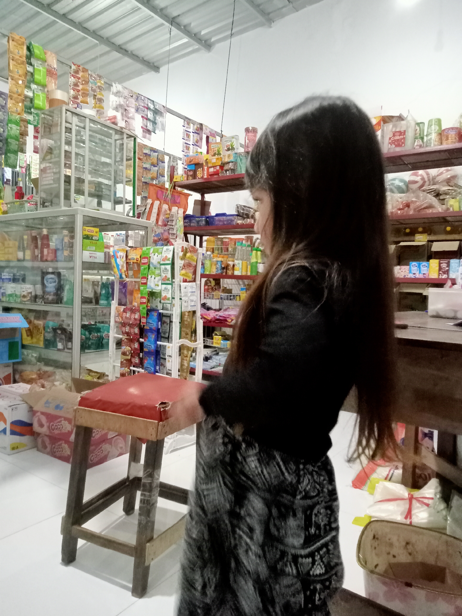 A young girl with long, dark hair stands in what appears to be a small convenience store or warung in Simo, Indonesia. She is wearing a dark, long-sleeved shirt and a patterned skirt. Her profile is visible as she looks to her right, towards shelves stocked with various products. The store is filled with merchandise, including packaged snacks, drinks, and household items displayed on shelves and hanging from the ceiling. There are glass display cases, a wooden stool with a red cushion in the foreground, and a counter to the right. The lighting is artificial, typical of an indoor retail space.