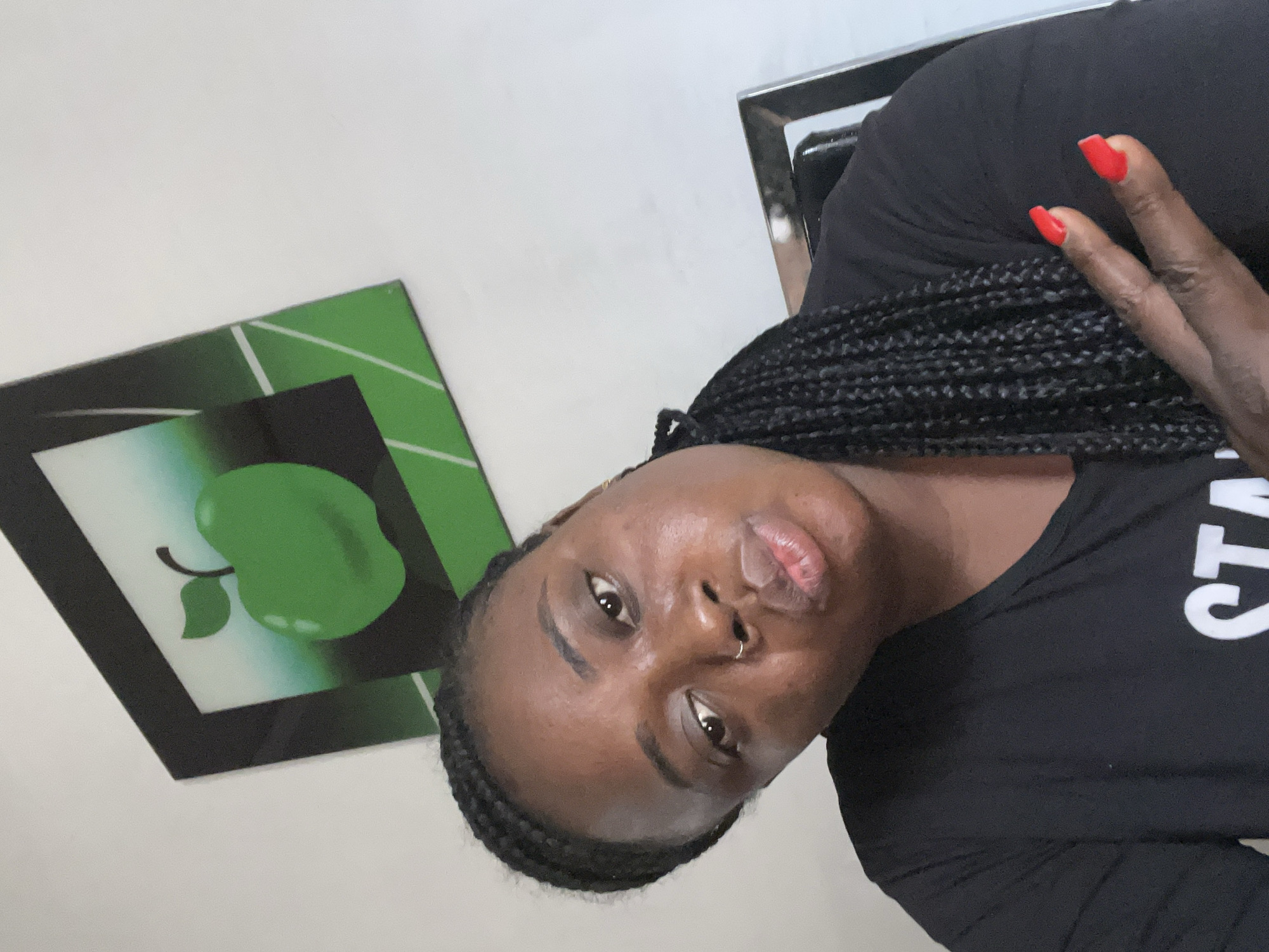 A close-up selfie captured indoors in Jos, Nigeria, features a person with dark skin and long black braided hair. The individual is looking directly at the camera with a neutral expression, exhibiting a slight pout on their lips. They have a septum nose ring. The person is wearing a black t-shirt, on which partial white text "STA" (likely part of "STAY" or similar) is visible. One hand is raised, displaying red, coffin-shaped nail polish and forming a two-finger gesture, resembling a peace sign.
The background consists of a light-colored wall. To the left, a framed picture or decorative panel is visible, featuring a bold green apple with a stem and leaf, set against a background of varying shades of green and black with white geometric lines. Part of a metallic, possibly chrome, frame of a chair or another piece of furniture is visible on the right side of the image. The lighting suggests it is daytime.