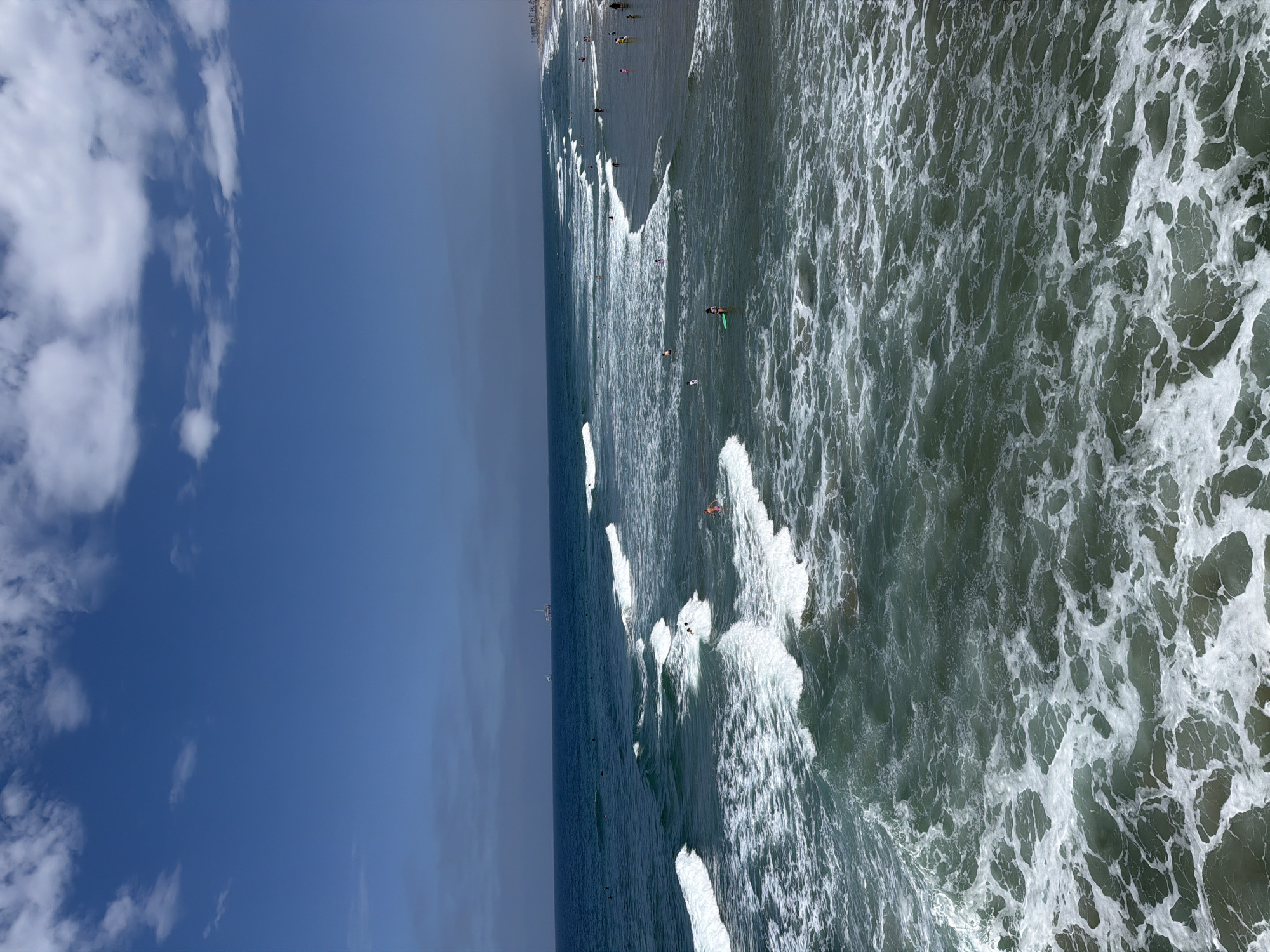 This is a wide-angle, elevated shot of the Pacific Ocean at Huntington Beach, California. The sky is a clear, bright blue with scattered white cumulus clouds. The ocean is a deep blue-green, with numerous white-capped waves rolling towards the shore. Several surfers are visible in the water, some riding waves and others paddling. A few people are wading or swimming near the shoreline, which is visible in the background as a sandy beach lined with palm trees and buildings. A large boat, possibly a barge or oil rig, is visible on the horizon. The overall scene depicts a sunny, active day at the beach.