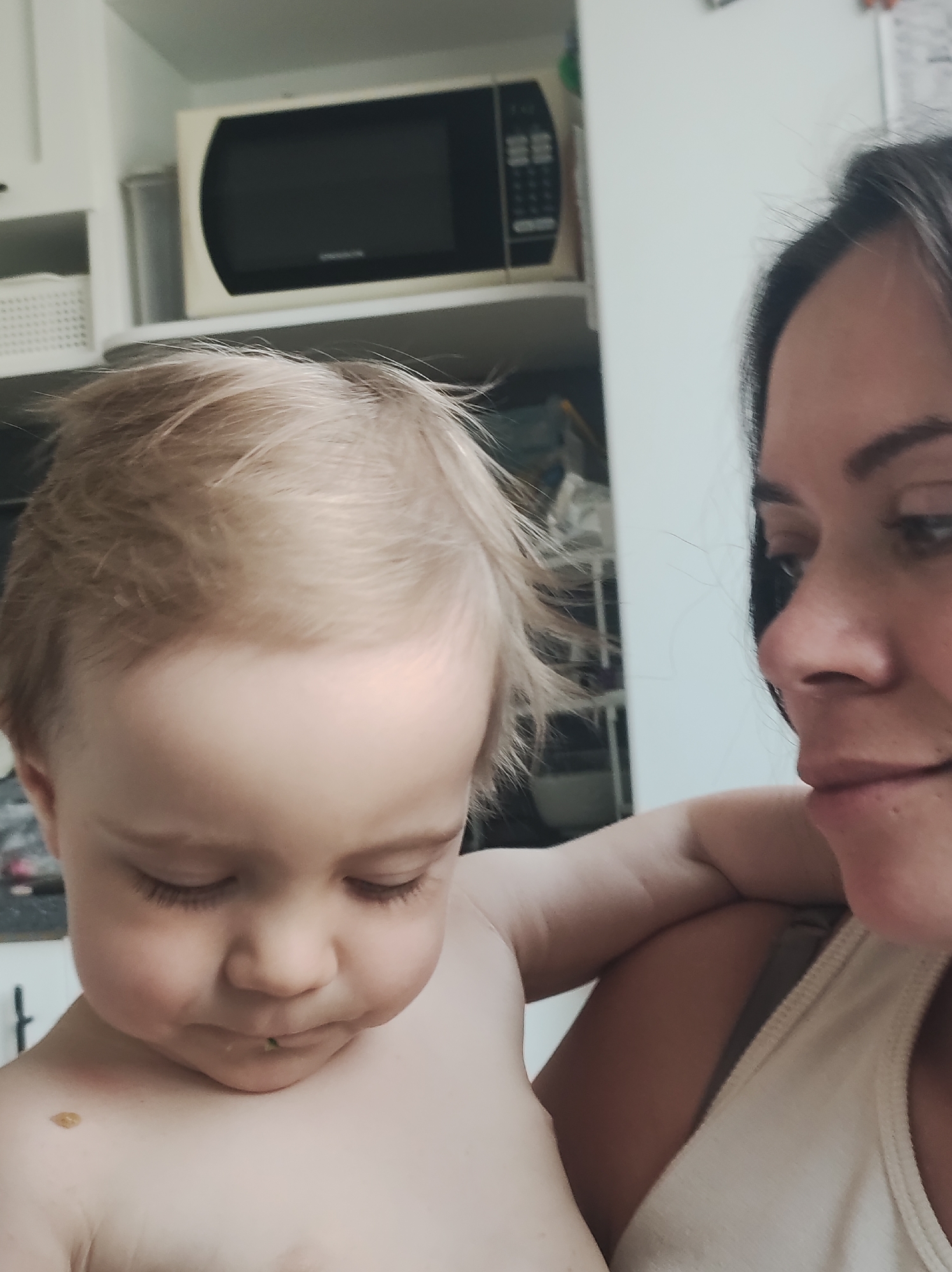 A fair-skinned child with light blonde hair, bare-chested with a small piece of green food on their lower lip, is held in the arms of a fair-skinned woman with dark hair and visible eyebrows. The woman is wearing a light-colored tank top. Behind them is a kitchen setting with a microwave oven above a cabinet. The exact location in the city of Chaikovsky, Russia is indeterminable from the visual information.