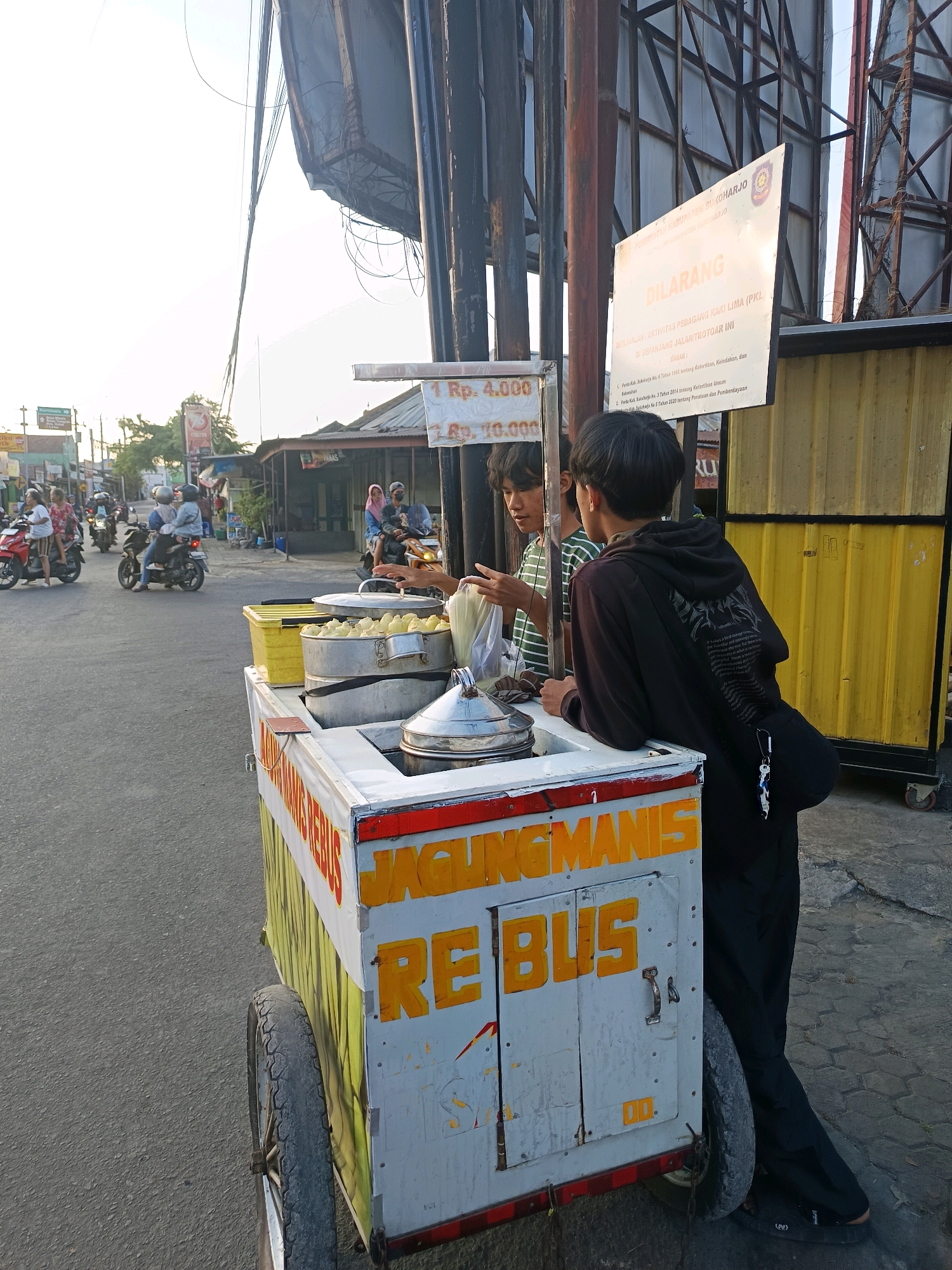 In Gatak, Indonesia, on a sunlit day, a street food vendor operates a cart laden with steamed corn. The cart, a white box with yellow and red trim, displays the words "JAGUNG MANIS REBUS" in large orange letters. Two young men stand near the cart; one, wearing a striped t-shirt, appears to be assisting the vendor, while the other, in a black hoodie, leans against the cart. In the background, the street bustles with motorbikes and pedestrians, and a sign in Indonesian warns against street vendors. The scene is a snapshot of everyday life in a Indonesian town.