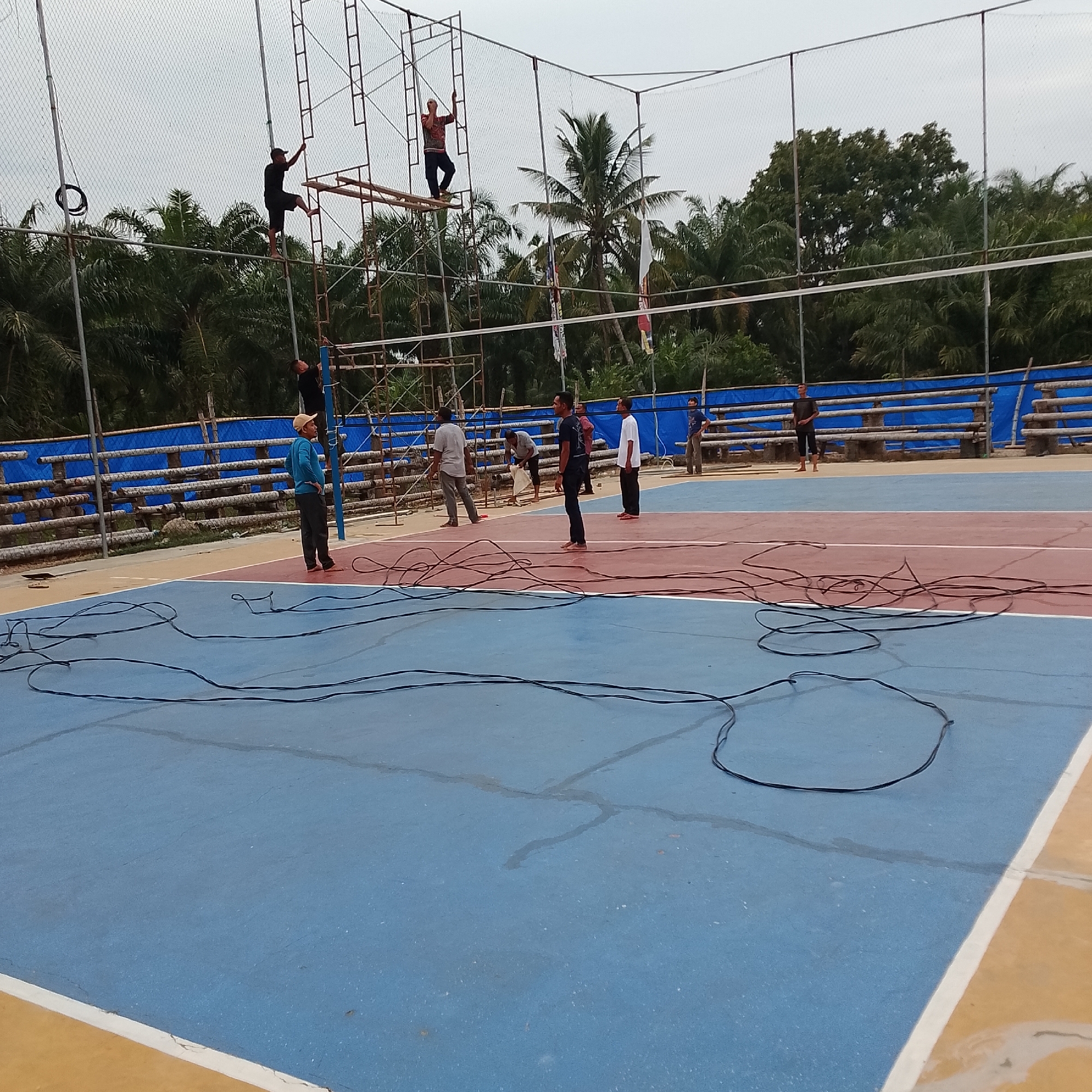 The image shows an outdoor volleyball court with a blue and red surface. In the foreground, black wires are spread across the court. Several people are present, some on the court and others on scaffolding near the net. Two individuals are visible on the scaffolding, appearing to be working. On the court, a person in a blue shirt and cap stands near the net, facing away from the camera. Other individuals are scattered on the court and in the background, some observing and others engaged in activity. The background features a blue tarp-covered barrier, wooden seating, and dense foliage with palm trees. The sky is overcast.