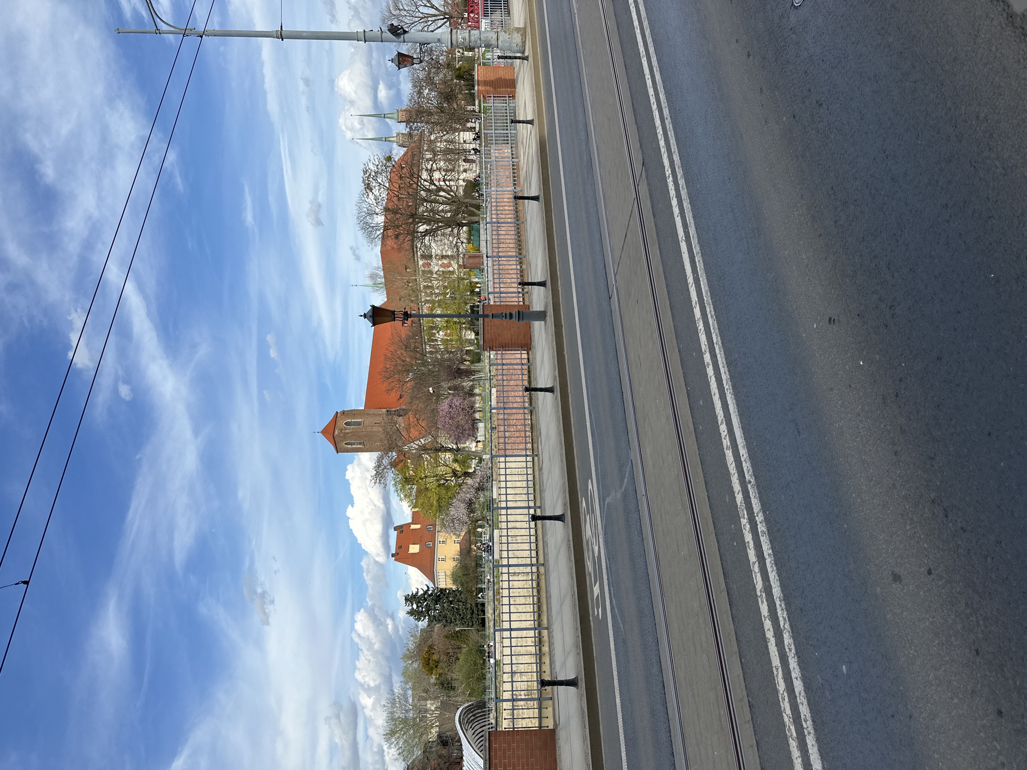 The image depicts a view from a high angle, possibly a bridge, overlooking a street scene in Wrocław, Poland. The scene is oriented vertically, with the road running lengthwise. A street with double white lines and tram tracks runs alongside a sidewalk bordered by a low brick and metal fence. Beyond the fence, the view includes various buildings, some of which are churches or other historical structures with distinctive red roofs and spires. The sky is visible above the buildings and is filled with clouds. The composition suggests a typical urban landscape within the city of Wrocław. There are no people visible.