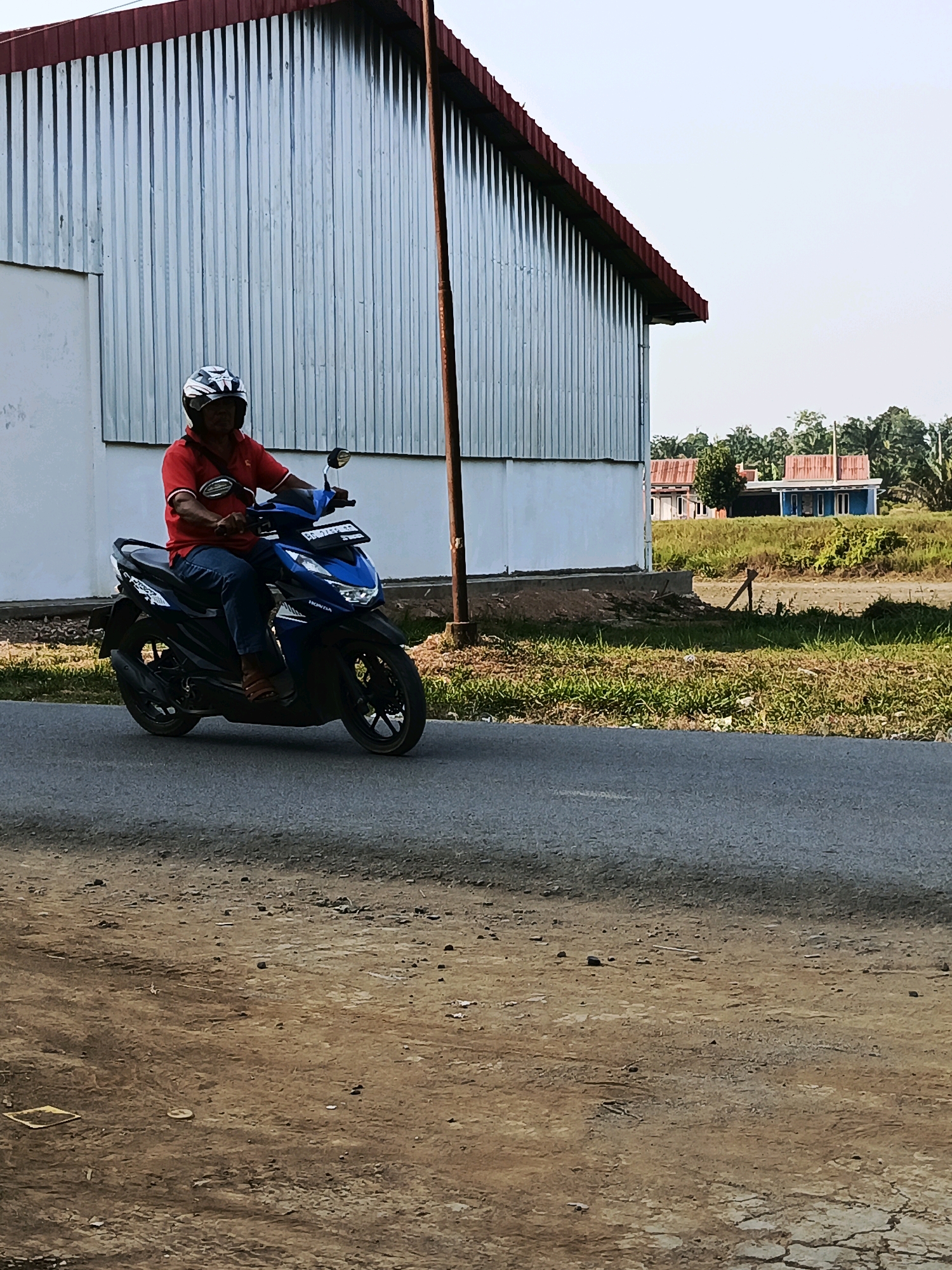 A man in a red shirt and blue jeans rides a blue Honda scooter on a paved road. He is wearing a white and black helmet. The scooter has a license plate that reads "BG 2157 CO". In the background, there is a long, corrugated metal building with a red roof. To the right of the building, there are two small houses with red roofs and a few trees. The ground in the foreground is a mix of dirt and gravel. The sky is clear and blue. This scene is located in Megang Sakti I, Indonesia.
