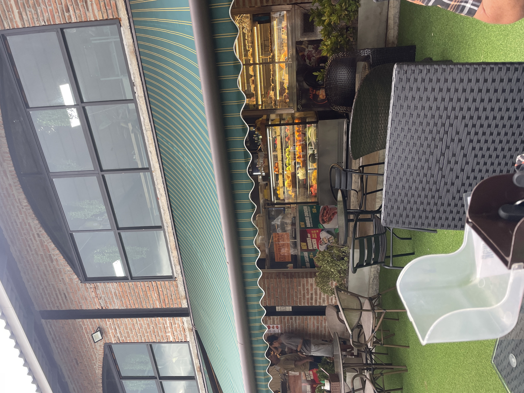 The image captures a scene in Choeng Thale, Thailand, showcasing the exterior of a bakery or restaurant. The building is constructed of red brick, featuring large glass windows with black frames. An awning with green and gold stripes covers the entrance, extending over a display case filled with various pastries and fruits. Outside seating is arranged on a patch of artificial green grass, including black rattan tables and chairs. Several chairs are empty, while a person is partially visible at one table. Display posters and menus, advertising items like "BANDARI CHICKEN" and "DARI SAUSAGE," are visible near the entrance.