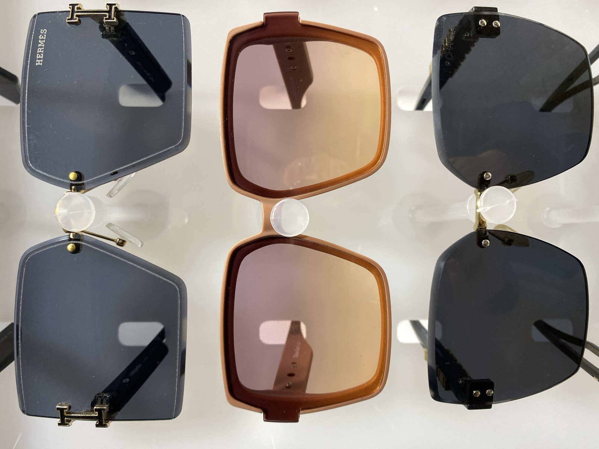 The image shows six pairs of sunglasses displayed on a white surface. The sunglasses are arranged in two rows of three. From left to right in the top row, the sunglasses are: a dark-lensed pair with the word "HERMES" on the lens and a gold H logo at the top; a brown-framed pair with gradient orange/brown lenses; and a dark-lensed pair with a gold logo at the top right. The bottom row mirrors the top row in arrangement and styles. The background is plain white. The location in Alanya, Turkey is not discernible from the image.