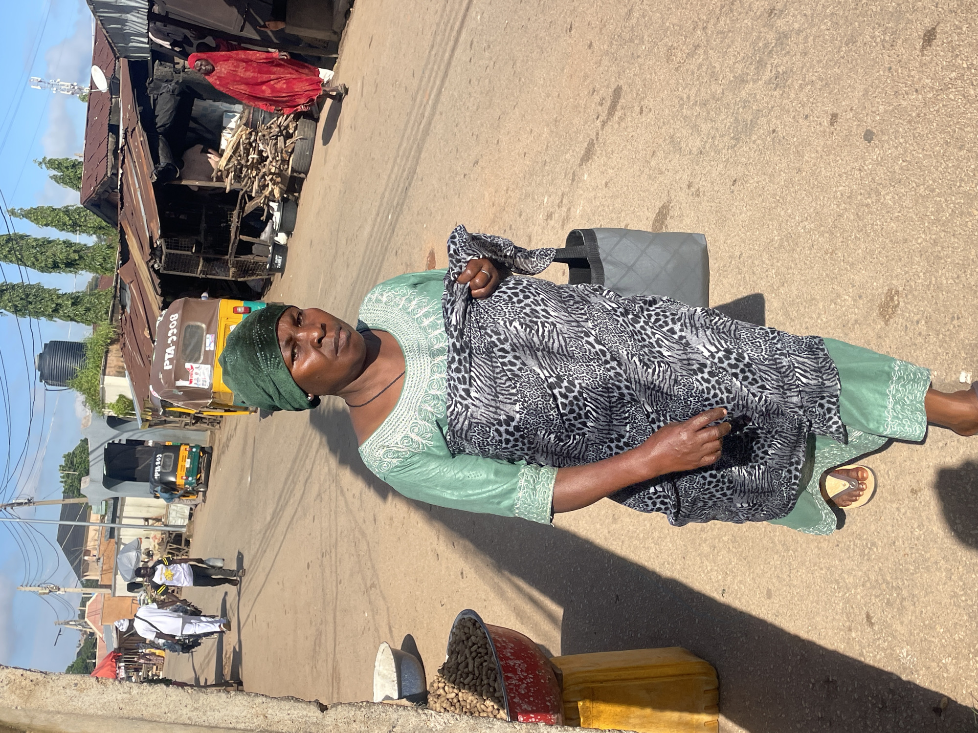 A medium shot captures a woman in a green headscarf and matching green patterned dress with a black and white animal print wrap over her right arm, standing on a dirt road in Jos, Nigeria. She holds a dark grey tote bag in her right hand. To her left, a red bowl filled with peanuts sits on a yellow stool. The road is lined with makeshift stalls constructed from corrugated metal, some with stacked firewood. In the background, a yellow and green auto-rickshaw with license plate "PTA 3308" is visible, as well as another person in a red garment walking away from the camera. The sky is blue with scattered clouds, and the scene is illuminated by sunlight, casting long shadows.
