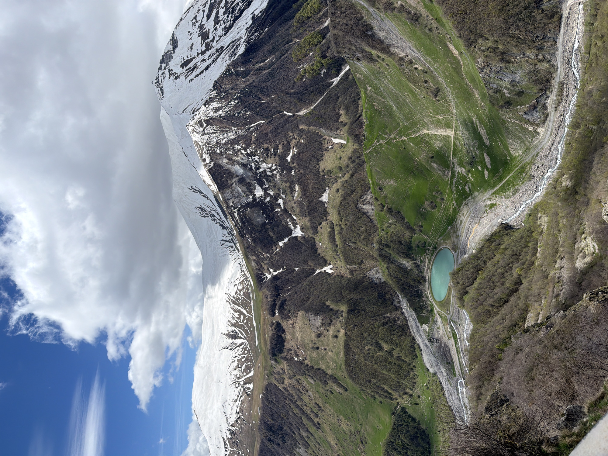 The image shows a high-angle view of a mountainous landscape, likely in the Gudauri region of Georgia. Dominating the left side is a large mountain partially covered in snow, contrasting with patches of exposed rock and vegetation. Clouds obscure the upper section of the mountain peak. To the right of the mountain, a steep valley descends, exhibiting varying shades of green, suggesting different types of vegetation. A small, turquoise-colored lake is nestled within the valley. A narrow river or stream runs through the valley, and a pathway or road snakes along the edge of the stream. The foreground has a few bare trees, possibly at the edge of a cliff. The sky is partly blue with light, wispy clouds.