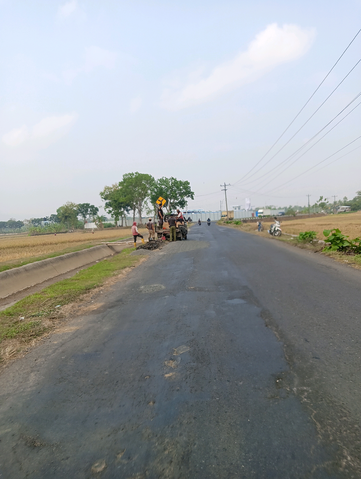The image depicts a roadside scene in Bokol, Indonesia, where road work is in progress. A group of construction workers are gathered around a damaged section of the asphalt road, with some actively engaged in repairs. They are using a small truck to transport materials, and tools are scattered around the work area. The road itself appears to be in disrepair, with visible potholes and uneven surfaces. The surrounding environment consists of a canal running alongside the road, dry agricultural fields, and trees. The sky is overcast, with a few clouds visible. Several motorcycles are present, indicating the mode of transportation in the area. The overall impression is one of ongoing infrastructure maintenance in a rural or semi-rural setting.