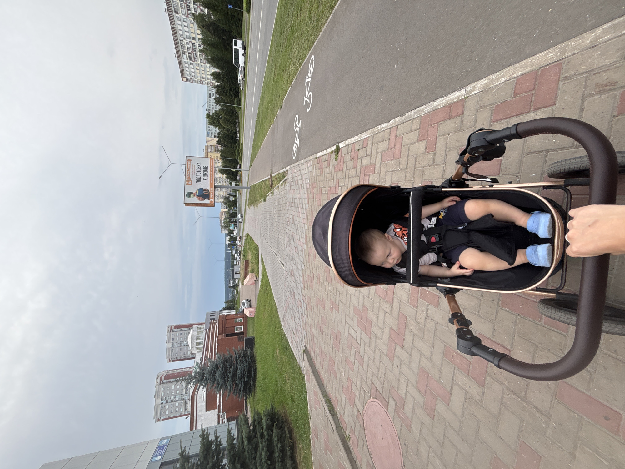 A first-person perspective shot captures a person pushing a baby in a stroller along a paved sidewalk. The baby, wearing a t-shirt and shorts with blue Crocs, sits facing forward in a black stroller with gold accents. To the right of the sidewalk, a paved bike lane is marked with white bicycle symbols. Beyond the bike lane is a road with a few cars and a white bus visible in the distance. A billboard advertising "Gorod Znaniy" (City of Knowledge) for school preparation is positioned on the left side of the road. In the background, multistory apartment buildings and trees line the street. The sky is overcast. The scene appears to be in an urban environment, likely Naberezhnye Chelny, Russia, given the Cyrillic text on the billboard.