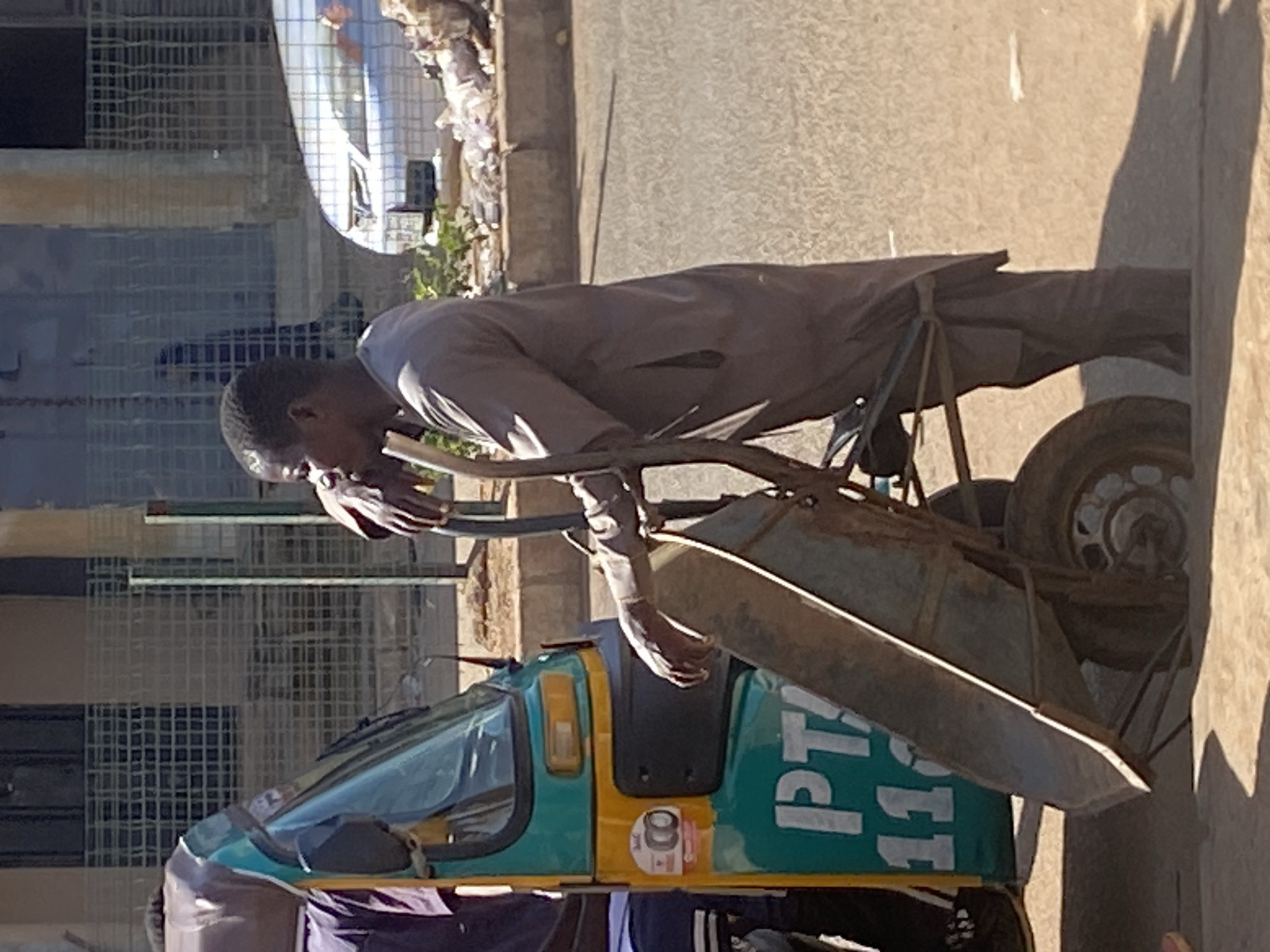 An outdoor scene in Bukuru, Nigeria features a man standing next to a colorful tuk-tuk, a three-wheeled vehicle used for transportation, and a wheelbarrow. The man, wearing a loose-fitting grey garment, is holding the handles of the wheelbarrow. The tuk-tuk is predominantly turquoise and yellow, with some signage on its side. The ground appears to be paved. In the background, a metallic fence is visible, and beyond that, a building and a silver car. The overall impression is of a street scene in a bustling urban environment.