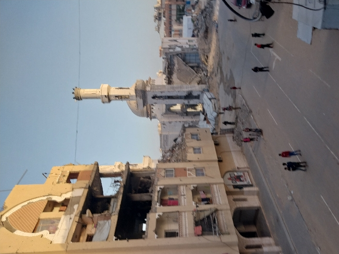 The image captures a scene of widespread urban devastation, likely from conflict or a major disaster, viewed from an elevated perspective. The photograph is oriented vertically.
In the center-left of the frame, a prominent, light-colored minaret stands tall amidst significant destruction. Its upper section, featuring a distinct, crown-like or turreted top, appears largely intact, though portions of its base or the attached mosque structure show damage, with exposed sections and debris. Behind the minaret, part of a dome is visible, also appearing to be damaged. The area immediately surrounding the mosque structure is reduced to extensive piles of rubble and debris, suggesting that nearby buildings have been completely destroyed.
In the bottom-left foreground, a multi-story building is severely damaged. Large sections of its exterior walls have collapsed or been blown out, exposing the interior rooms, twisted rebar, and structural elements. The building exhibits a mix of concrete and what appears to be decorative architectural details, now broken and fragmented. Inside the exposed rooms, various remnants are visible, indicating prior occupancy.
On the right side of the image, a wide, paved road stretches from the foreground into the distance. White lane markings are visible on its surface. Numerous individuals, appearing to be adults and possibly some children, are walking along the road. They are dressed in casual attire, with some wearing red or dark jackets. They are scattered across the width of the road, moving in various directions, seemingly navigating the destroyed environment.
The background, above the minaret and extending beyond the road, reveals more heavily damaged or completely destroyed buildings and vast expanses of rubble, indicating the widespread nature of the devastation across the urban landscape.
The sky, visible in the upper-left portion of the image, is clear and light blue, suggesting daytime and fair weather. The lighting appears soft, possibly indicating early morning or late afternoon. No discernible text is visible within the image. The overall impression is one of a city grappling with the aftermath of a destructive event, with inhabitants moving through the ruins.