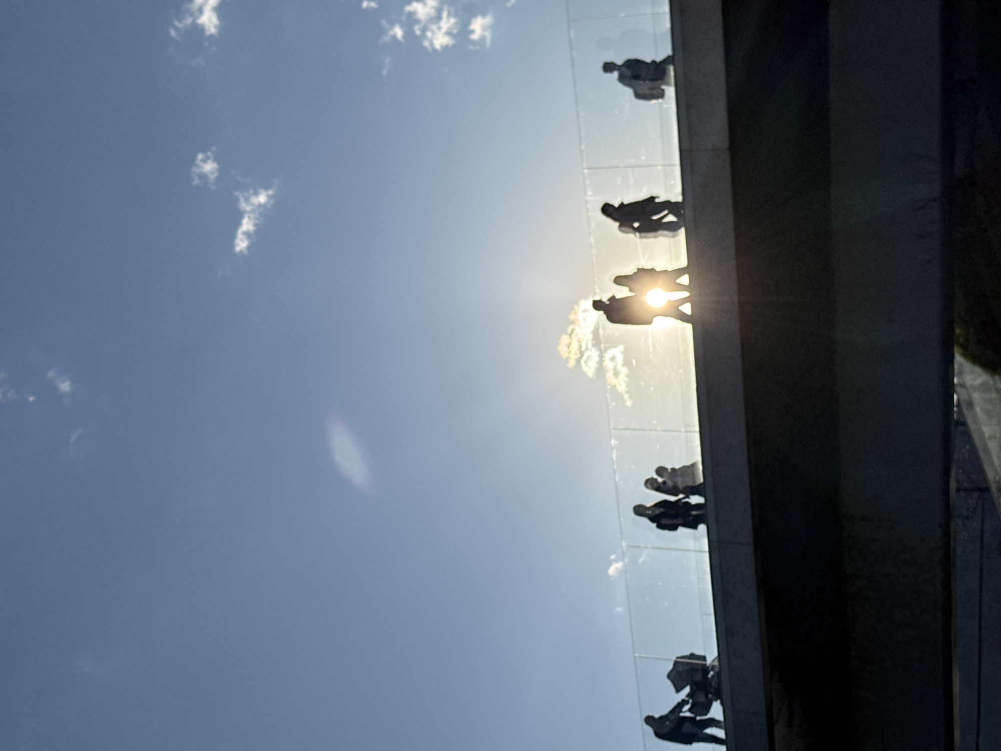 The image is a low-angle shot showing the "Soaring Bridge" at Zaryadye Park in Moscow, Russia. Silhouetted figures of people are walking on the bridge, which is suspended over the Moscow River. The sun is directly behind some of the figures, creating a strong backlighting effect. A clear, blue sky with scattered clouds forms the background. The edge of the bridge structure, appearing as a dark, vertical element, frames the scene on the right.