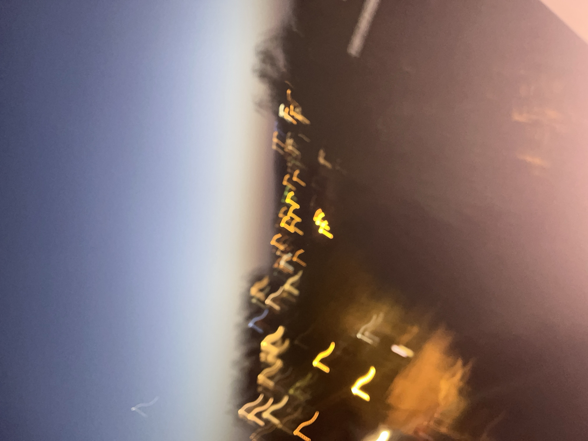 The image shows a blurred cityscape at night, with streaks of yellow and orange light likely representing streetlights or vehicle lights. The sky is visible on the left, transitioning from dark blue to a lighter hue near the horizon. The location appears to be an urban area.