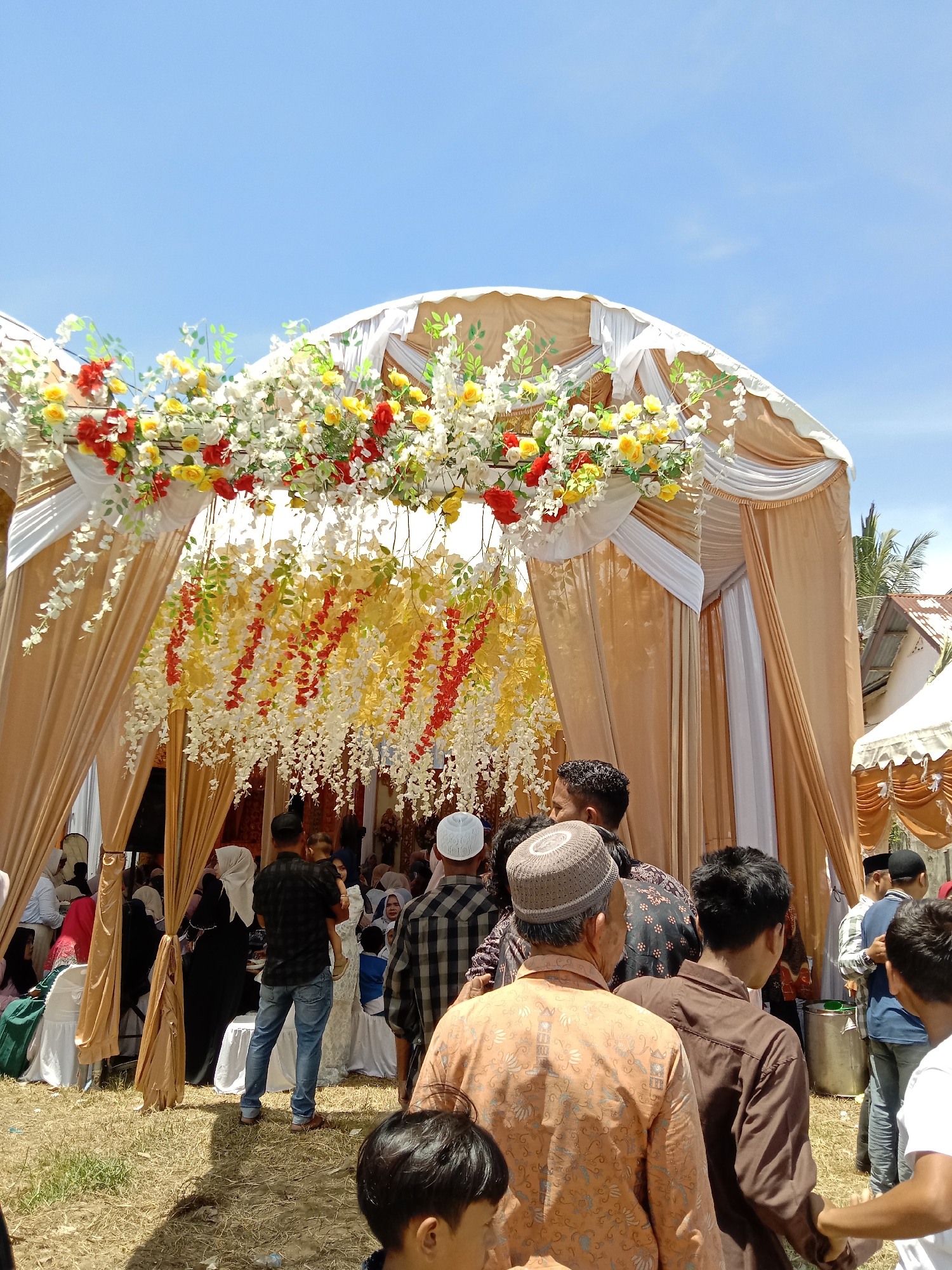 The image captures an outdoor event, possibly a wedding celebration, in Lhokseumawe, Indonesia. A large tent-like structure serves as the main focal point, adorned with a profusion of white, yellow, and red floral decorations. The structure is draped with tan and white fabric, creating a formal and festive atmosphere.
A crowd of people is gathered near the entrance of the tent, many wearing traditional Muslim clothing. Some individuals are positioned inside the tent, where seating arrangements can be seen. The foreground shows the backs of several people, suggesting a gathering or procession.
The environment appears to be a grassy area, possibly a field or yard. The sky above is clear and blue. In the background, a glimpse of a building and a small tent is visible. The lighting suggests a sunny day.