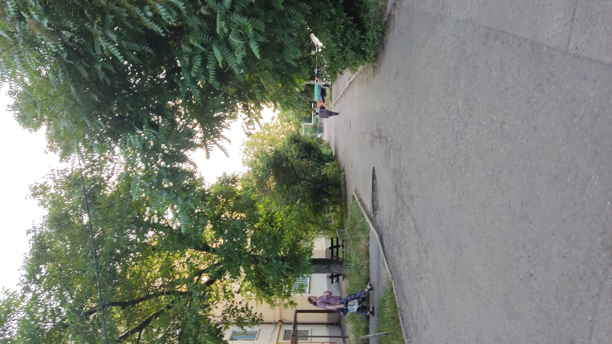 A woman and a child are walking on a paved path in front of a pale yellow building with some trees and bushes. The woman is wearing a purple t-shirt and patterned pants, and the child is wearing a white shirt and blue shorts. The path leads to a street with a few parked cars and more trees. The sky is bright and overcast, suggesting it is daytime. The scene appears to be in a residential area of Krasnodar, Russia, likely in front of an apartment building.