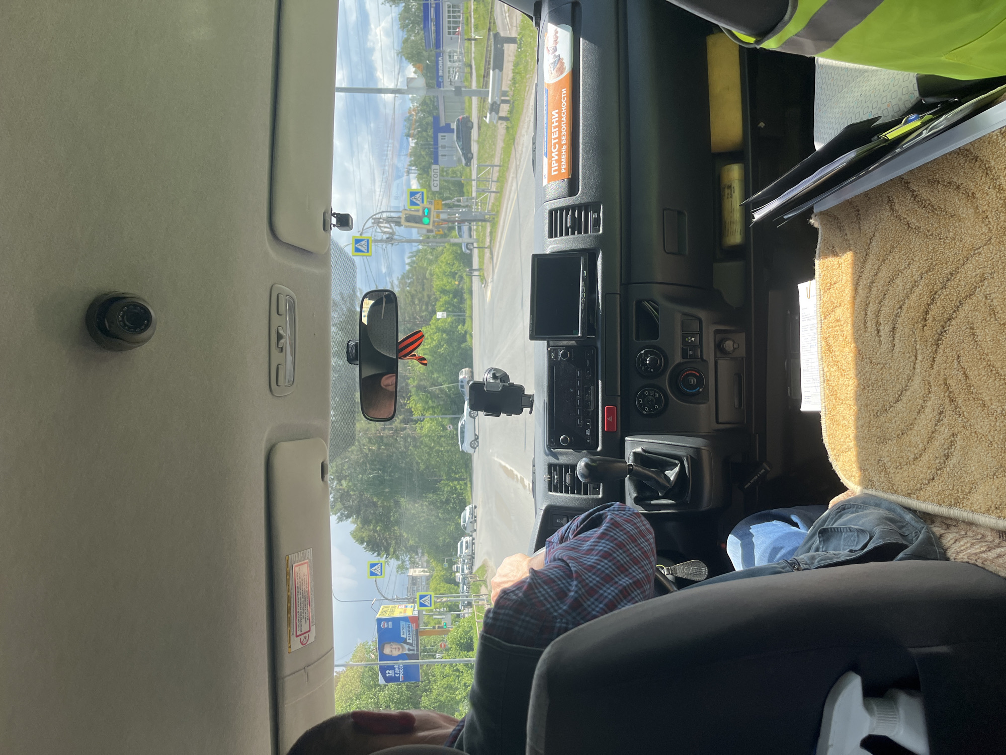 The image depicts the interior of a vehicle, specifically the front cabin. The driver's seat is partially visible, with an individual wearing a plaid shirt seated in the driver's seat. The dashboard features a central console with a display screen and various buttons and controls. A rearview mirror is mounted in the center. The vehicle is on a road with several pedestrian crossings. In the background, there is a building. The image indicates the location is Chaikovsky, Russia.