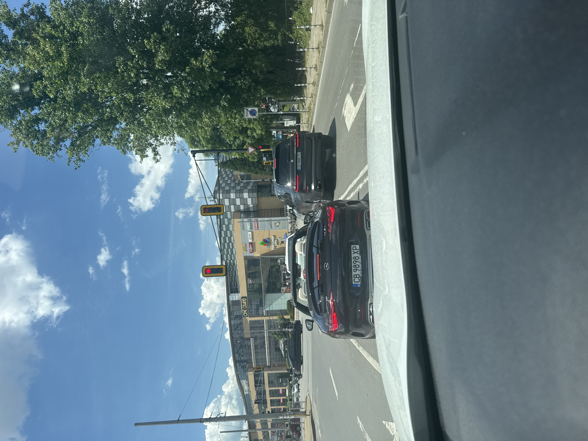 The image shows a view from inside a vehicle in Sofia, Bulgaria. The car is stopped in traffic at a red light. To the left is a shopping center complex called "Park Center Sofia." The photo captures two cars ahead: a Mercedes with license plate "CB 9898 XP" and a BMW with license plate "CB 9480 TC." The sky is blue with scattered white clouds, and there are trees lining the street. A speed limit sign of 30 km/h is visible.