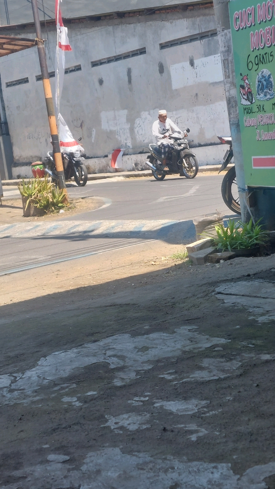 A man wearing a white cap and shirt rides a motorcycle on a paved road. The man is in the process of turning at an intersection. In the background, there is a concrete wall with horizontal slits, and a tall pole with flags. To the right of the man, there is a green sign with text in Indonesian. The scene appears to be in Sidoarjo, Indonesia, as indicated by the text on the green sign.