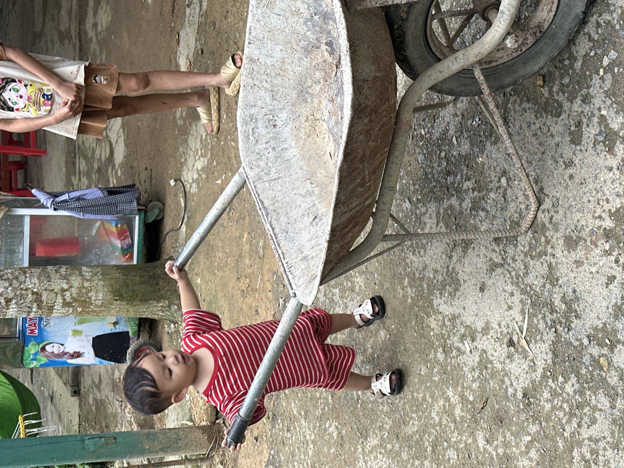 In Cổ Lũng Commune, Vietnam, a young boy wearing a red and white striped t-shirt and shorts, and white sandals, pushes a rusty metal wheelbarrow. He is positioned to the left of the wheelbarrow, his hands gripping the handles. To the right of the boy and partially obscured by the wheelbarrow, a second person, likely a child, stands wearing a graphic t-shirt and brown shorts, with light-colored sandals on their feet. The ground is a rough, uneven surface of dirt and gravel. In the background, a tree trunk is visible, along with a partially enclosed structure that appears to be a cart or stall with a poster featuring a woman. A green hammock is also visible in the upper left corner of the image.