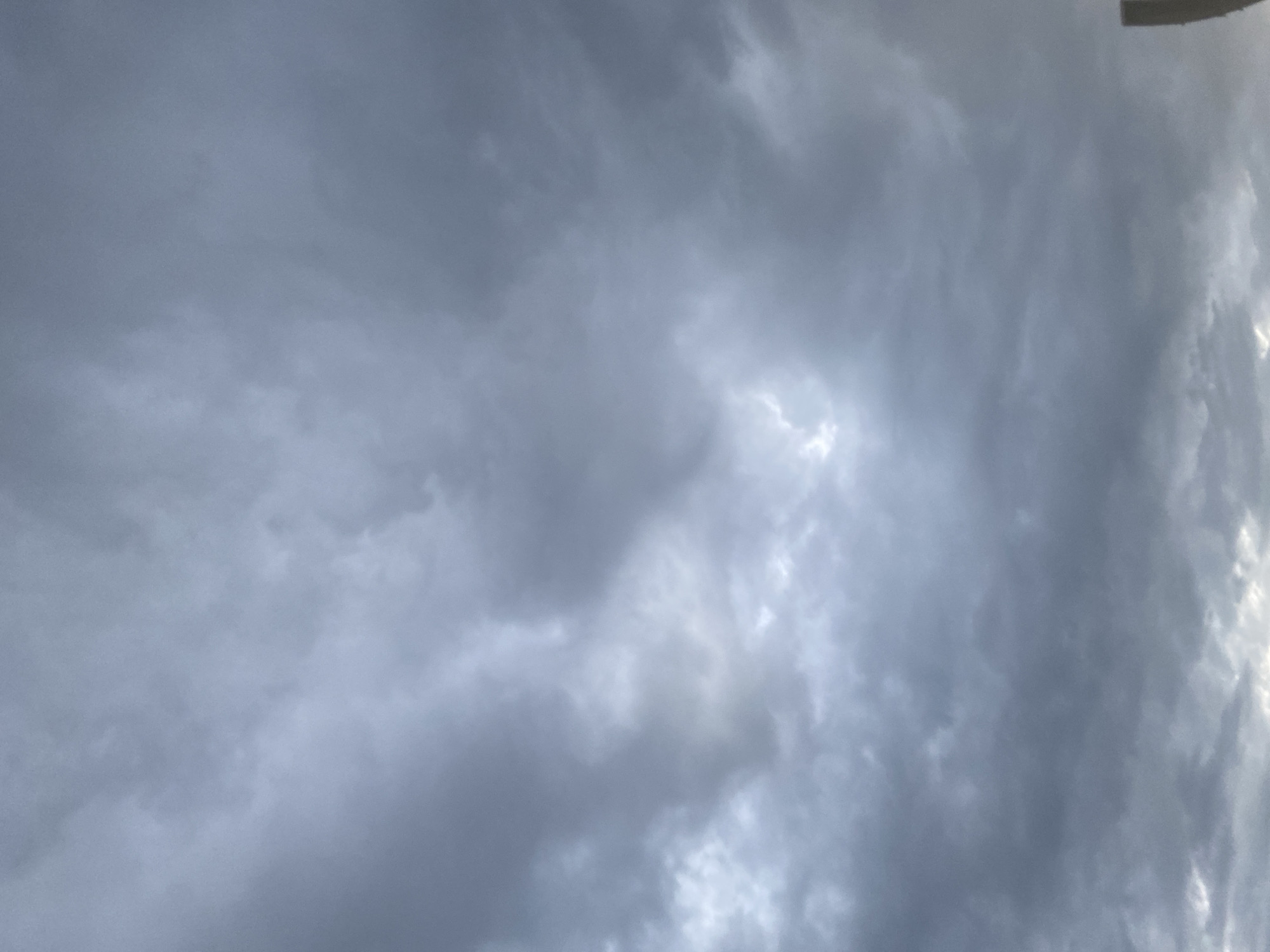 The image is a close-up of a cloudy sky. The clouds are dark gray and appear dense, suggesting overcast or approaching stormy weather. There is a small patch of lighter sky visible in the upper center of the image. A thin, vertical, light brown object, possibly a pole or antenna, is visible on the far right edge of the frame, extending from the bottom upwards. The overall lighting is dim and diffused. The image does not contain any people or explicit signs of urban activity within the frame, and therefore no specific location in Gurlan, Uzbekistan can be definitively identified from the visual content alone.