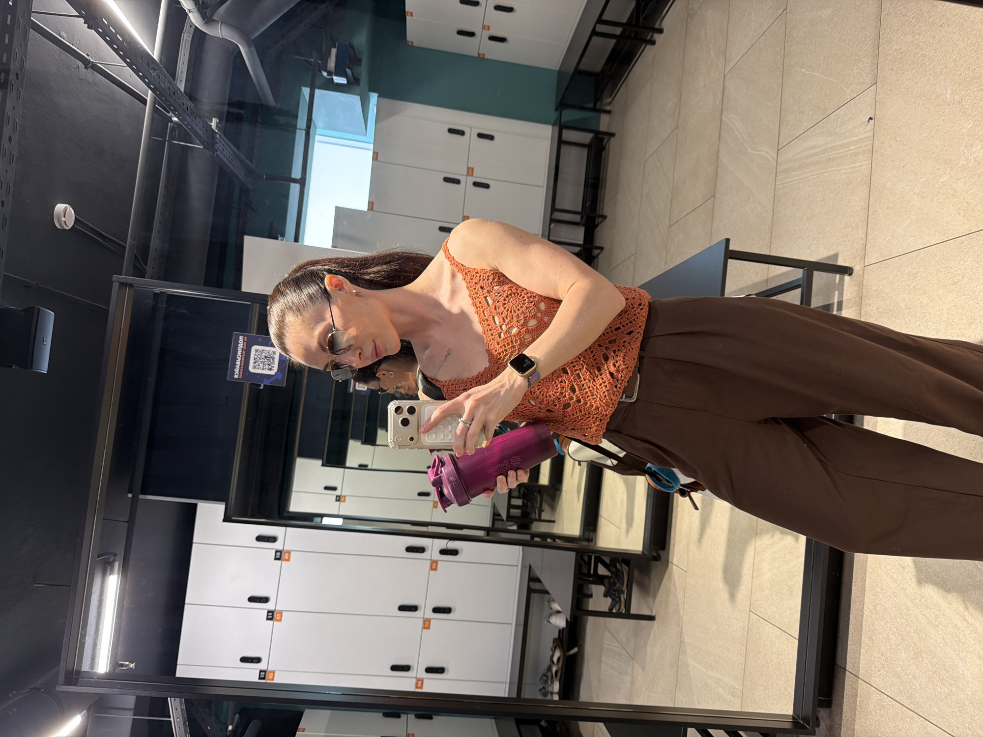 A woman with dark hair and sunglasses poses for a mirror selfie in what appears to be a locker room setting. She's holding a purple shaker bottle and her phone. She is wearing an orange crocheted top and brown trousers. The mirror reflects a row of white lockers and some storage racks. A QR code is visible on the mirror. The locker room appears to be in a modern facility. The location appears to be in Perm, Russia, as indicated by a QR code on the mirror that links to a Russian-language webpage.