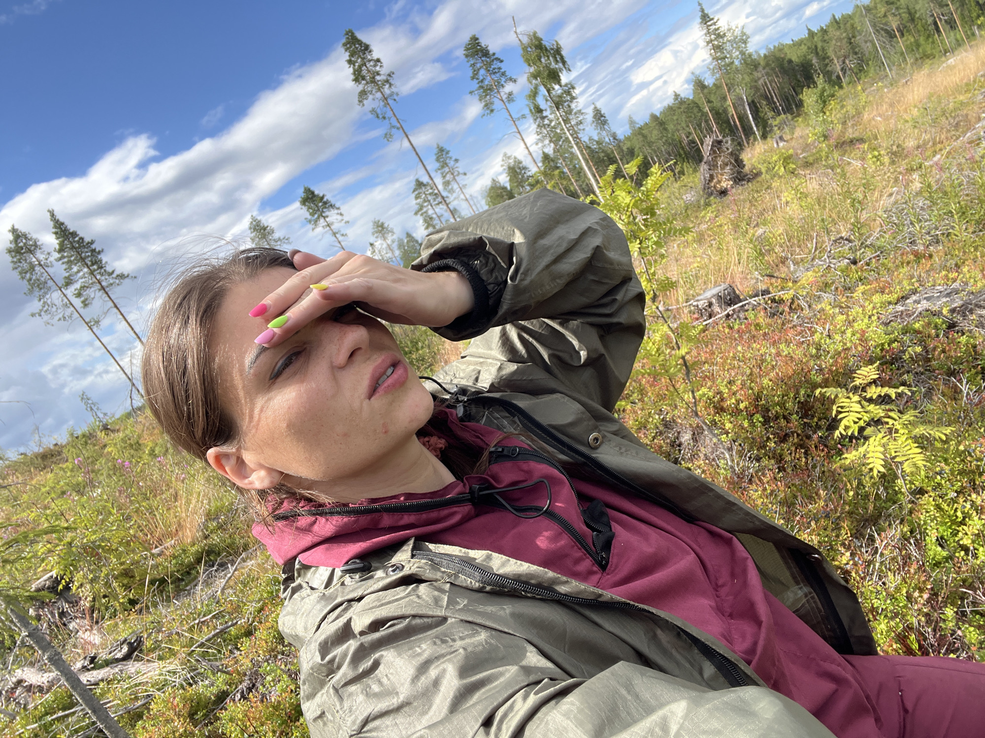 A woman is taking a selfie in a wooded, mountainous area. She is wearing a green jacket over a burgundy hoodie. Her hand is covering her eyes, and her nails are painted in bright colors. The background shows trees and undergrowth, with a cloudy blue sky above. The location is the city of Tolloreka, Russia.