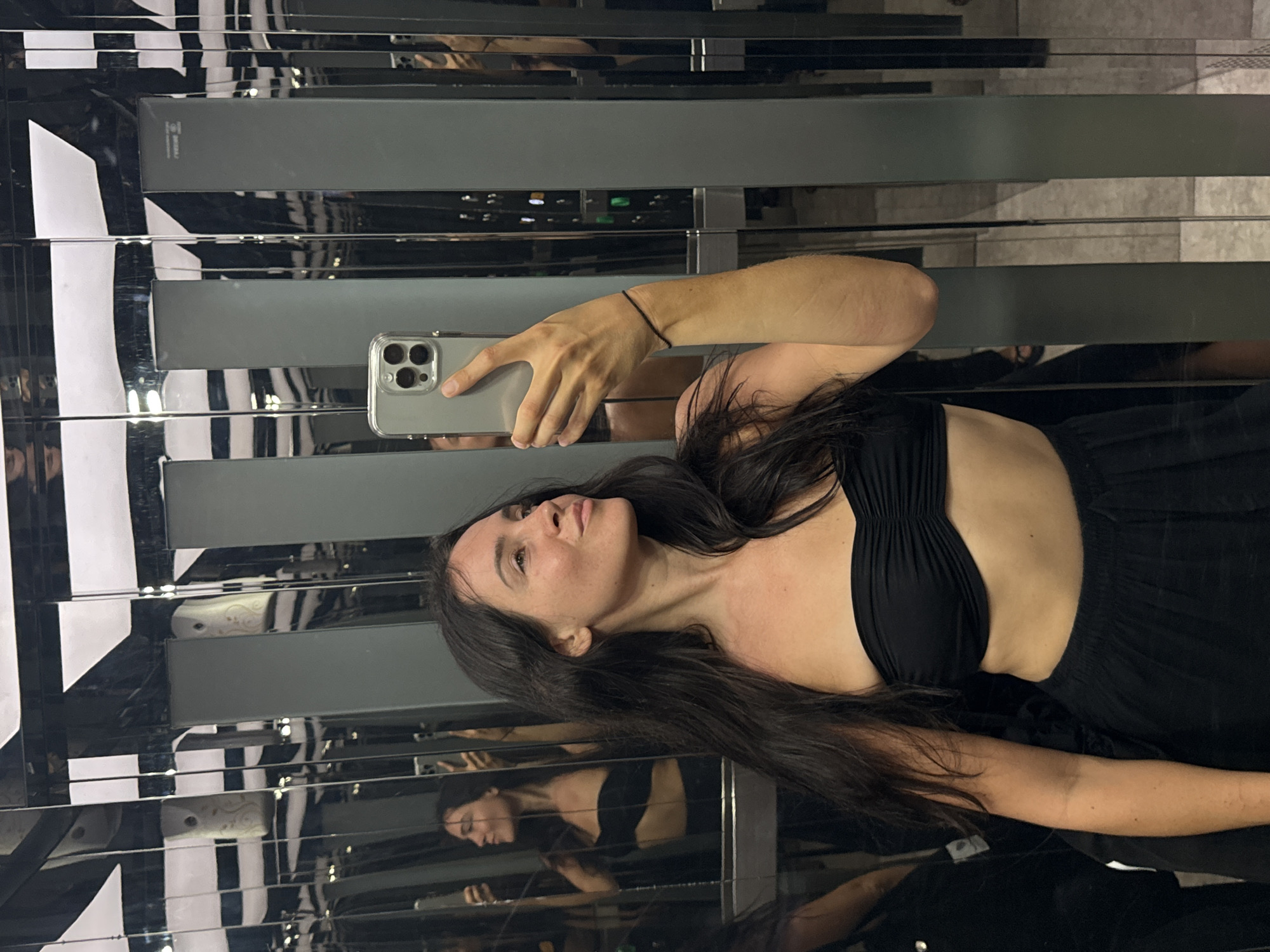 A person is taking a selfie in a mirrored elevator. The person, with long dark hair, is wearing a black bikini top and black pants. They are holding a smartphone in their right hand, which is visible in the reflection. The elevator walls are lined with mirrors and gray panels, creating a disorienting effect with multiple reflections. The overall scene is indoors and appears to be during the day, given the lighting. The person's expression is relaxed, and they seem to be posing for the photo. There are no visible text elements in the image. The location context of North Kuta, Indonesia, does not provide specific visual cues within the image itself.