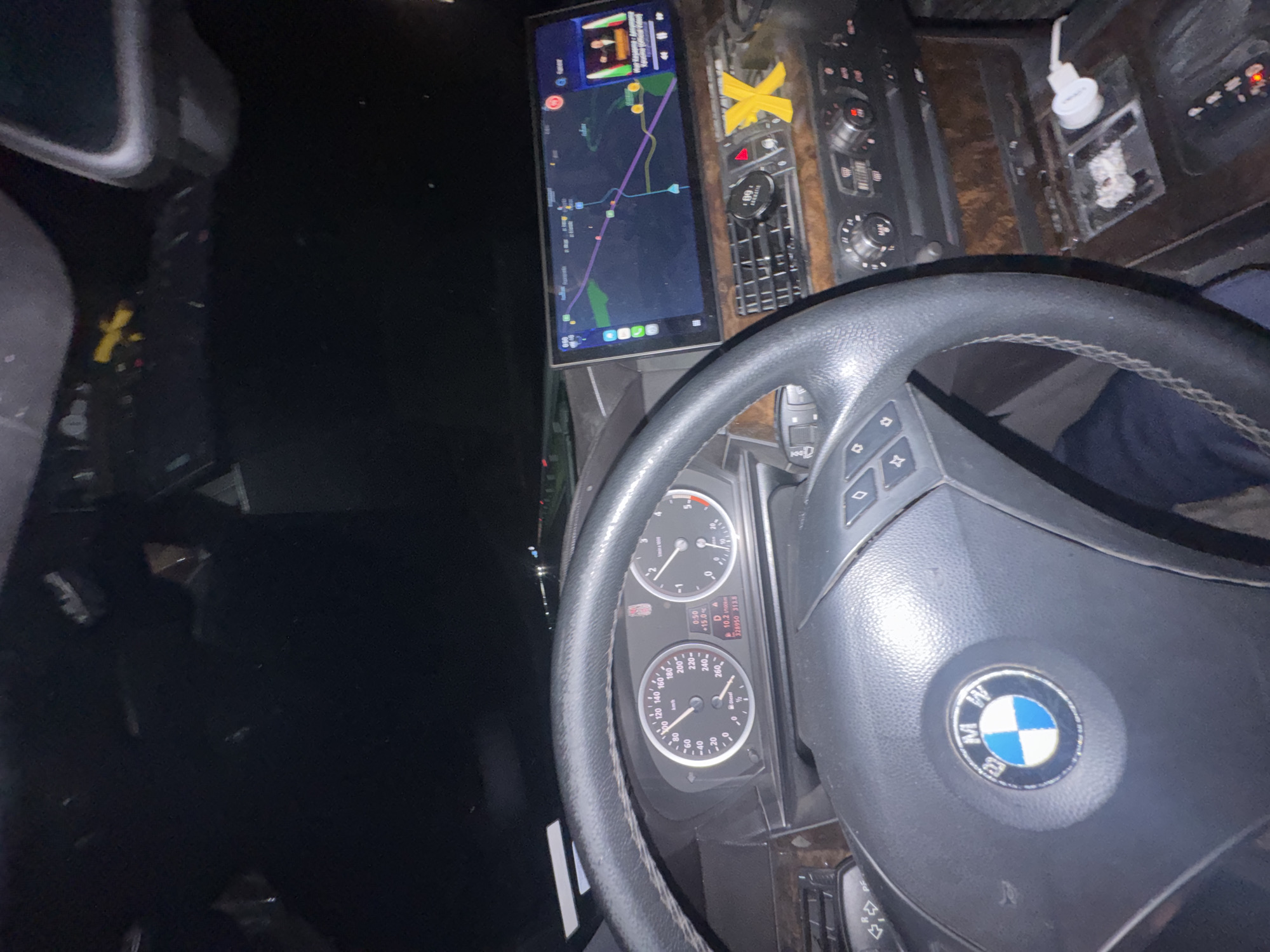 The image shows the interior of a BMW car, focusing on the driver's perspective. The steering wheel, instrument panel, and a screen displaying a navigation map are visible. The odometer reading is 328950 km. The navigation screen shows a route with a purple line, suggesting that the vehicle is potentially in transit. The location visible on the navigation map is in Mogila, Bulgaria. The environment appears dark, indicating that the image was taken at night or in a dimly lit area. The dashboard shows various controls and buttons, indicating a complex interface of the car.