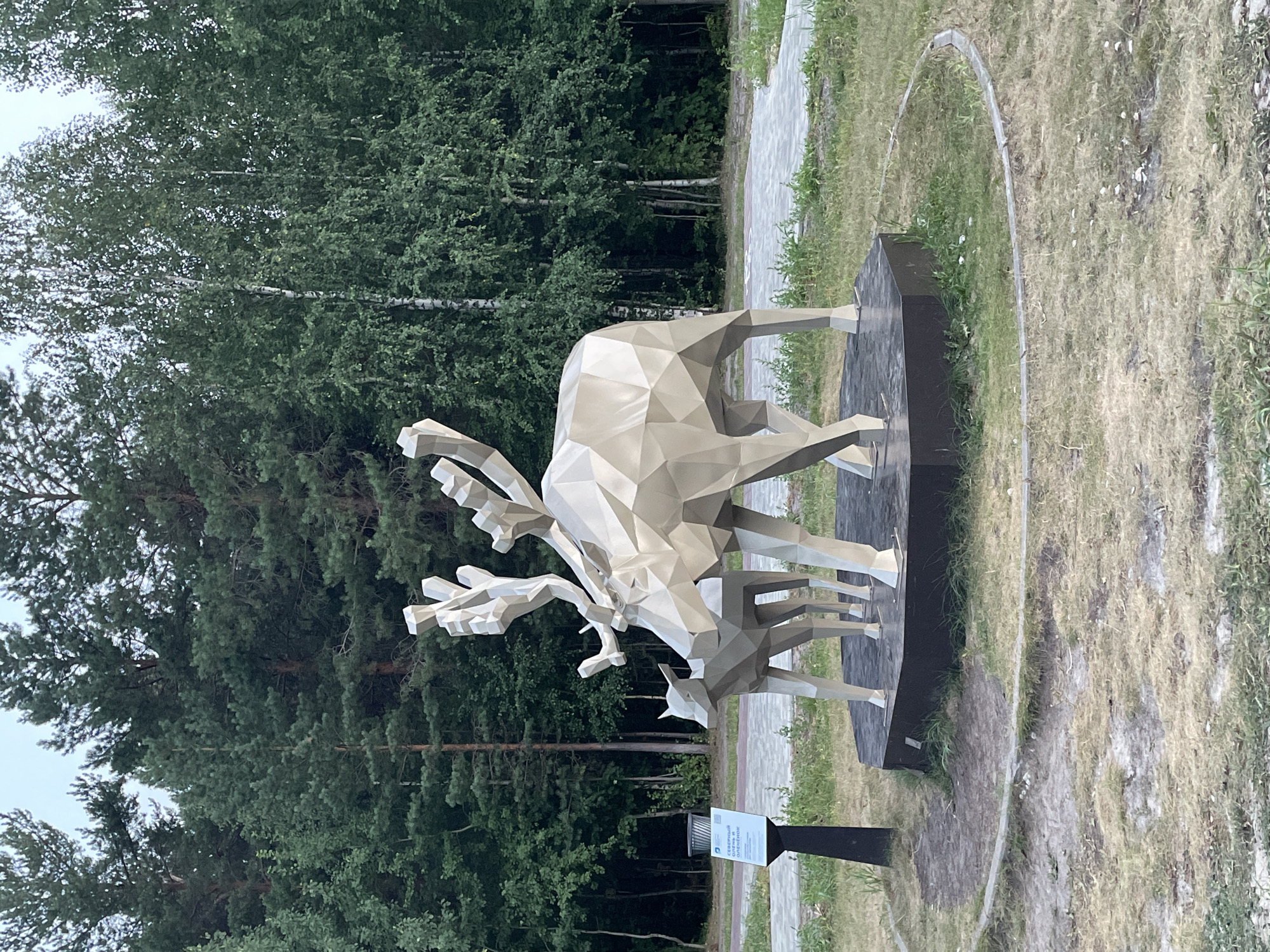 This media file shows a low-poly sculpture of a moose and calf, likely made of metal, in an outdoor setting. The sculpture is a light, reflective color, with angular facets creating its form. It is mounted on a dark, rectangular base.
The sculpture is situated in a park-like environment with a paved path visible behind it. To the left and background, there is a dense forest with tall, green trees. The ground around the sculpture is covered in grass, some of which appears dry and patchy, suggesting it might be summer or early autumn. A light gray, curved border runs around the area where the sculpture is placed.
There is no visible activity taking place; the scene is static and focused on the artwork. The time of day appears to be daytime, with diffused light suggesting an overcast sky or a shaded area.
A small sign is visible near the base of the sculpture. The visible text on the sign appears to be in Cyrillic script and reads: "СИМВОЛ ОЛЕНЬ ОБЕСПЕЧЕНИЯ". This translates to "Symbol of the Deer of Provision" or "Deer Symbol of Supply".
The location context provided is Surgut, Russia, which aligns with the presence of coniferous forests and the use of Cyrillic text. The overall impression is of a public art installation in a natural or semi-natural environment.