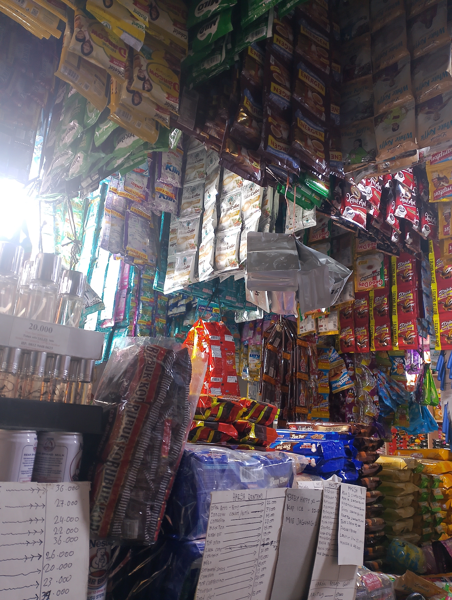 This image depicts the interior of a small store in Kedaidurian, Indonesia, likely a warung or mini-mart. The store is densely packed with various food and beverage products, primarily packaged goods such as instant coffee, milk powder, snacks, and instant noodles. These items are displayed in a disorganized manner, hanging from the ceiling, stacked on shelves, and piled on the floor.

In the foreground, there's a close-up of a price list written on white paper, detailing various items and their prices. To the left, there are rows of small spray bottles, possibly perfumes or essential oils, and two cans with labels that read "BEAR BRAND MILK". In the center and right of the image, the shelves are filled with a variety of colorful packaging. Prominent brands include "Energen", "Good Day", and "Kapal Api". Some products are arranged in clear plastic bags.

The lighting suggests an indoor setting, with a bright light source from the left, possibly a window or an open door, casting shadows and creating a slightly overexposed area. The overall impression is one of a busy, functional retail space catering to local needs, with a limited amount of space and a wide array of products. There are no people visible in the frame. The environment appears to be somewhat cluttered due to the sheer volume of merchandise.