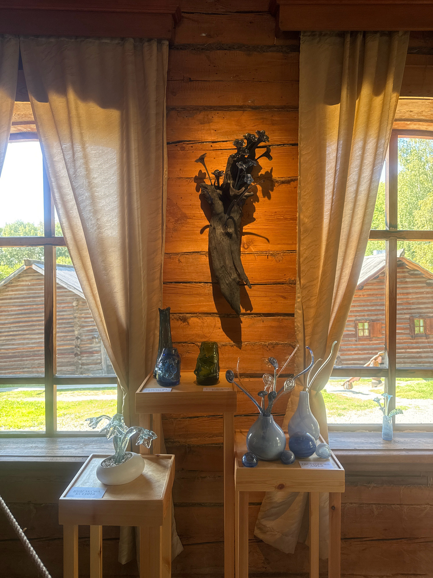 The image depicts an indoor scene within a rustic wooden building, likely part of the Taltsy Museum of Architecture and Ethnography in Irkutsk, Russia. Sunlight streams through two large windows, illuminating the wooden log walls and casting shadows.
In the foreground, two light-colored wooden display stands showcase various glass art pieces. The left stand holds a white, oval-shaped pot containing a clear glass plant-like sculpture. A label is visible on the front of this stand.
The right stand features a collection of glass objects. There are two vases, one light blue and one a lighter, pale blue. Several spherical glass objects in shades of blue are placed around the vases. In front of these, a label is also present.
Behind the display stands, mounted on the wooden wall, is a dark, sculptural piece that appears to be made of wood and dark metal, possibly depicting stylized flowers or branches.
The windows offer a view of the exterior, revealing two traditional wooden log houses (izbas) with peaked roofs, surrounded by greenery and sunlight. The setting suggests a museum or historical exhibit focused on traditional Russian rural life.
There are no people clearly visible in the interior of the scene. The overall impression is one of display and presentation of artistic creations within a historical, architectural context.