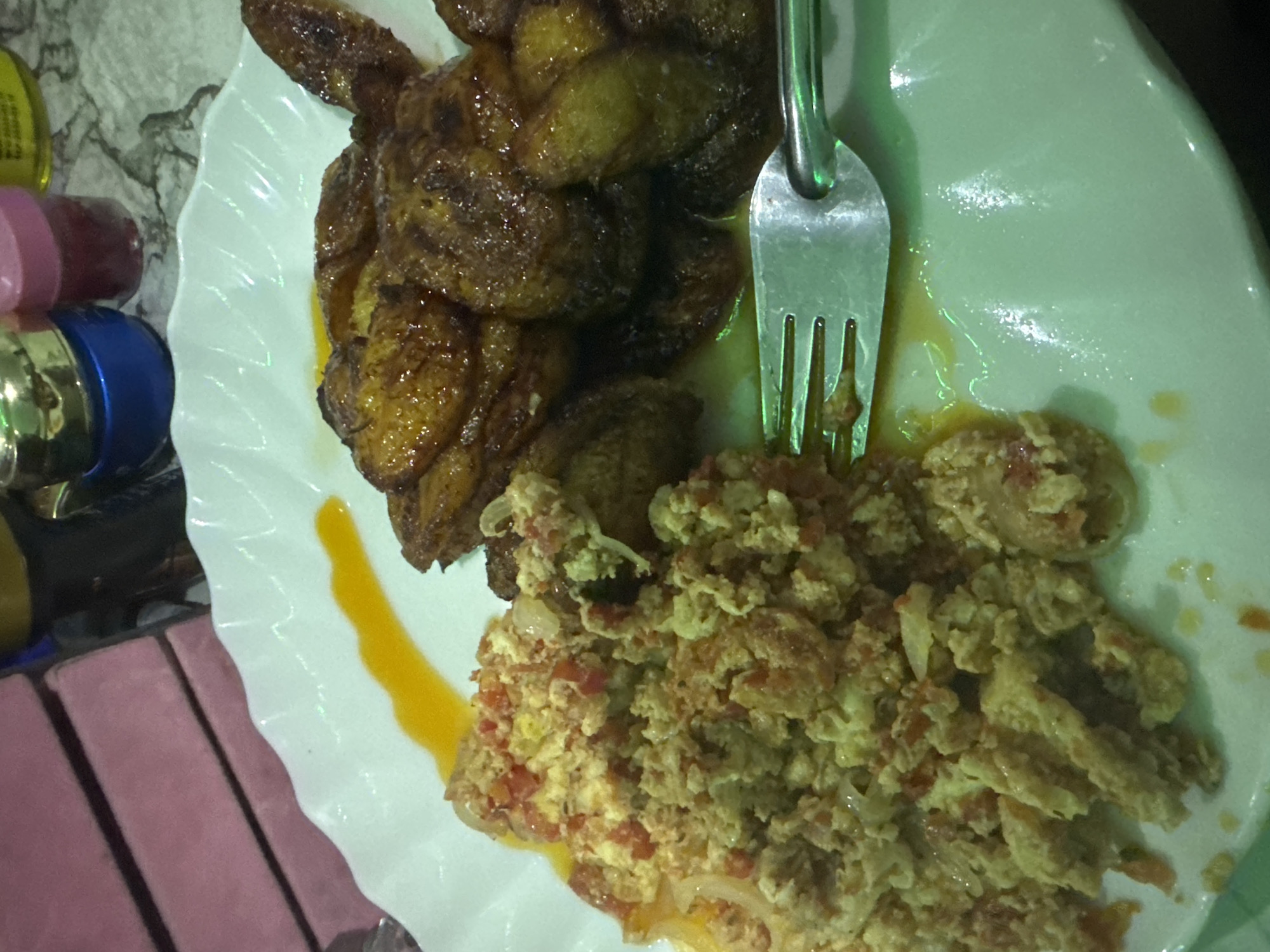 The image depicts a meal served on a white plate. The meal consists of fried plantains, which are piled in the upper left portion of the plate, and scrambled eggs with tomatoes, onions, and other vegetables, arranged in the lower right portion. A metal fork is partially embedded within the scrambled eggs and plantains. A streak of yellow sauce or oil is visible on the plate between the plantains and the eggs. In the background, there are blurred objects including small containers, possibly holding spices or condiments. To the side, there is a pink table or surface. This appears to be a homemade meal, likely being consumed in a residential setting in Asipa, Nigeria.
